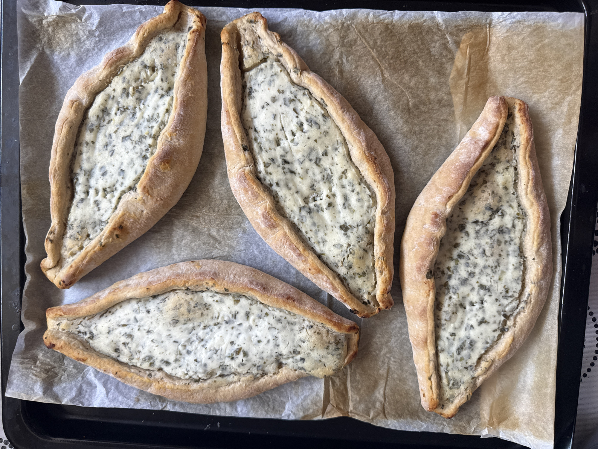 The image shows four baked boat-shaped pastries on a baking sheet lined with parchment paper. The pastries have a crusty, bread-like edge and are filled with a creamy, herb-flecked filling. The pastries are arranged closely together on the sheet. The scene is set on a black baking sheet, which is placed on a surface.