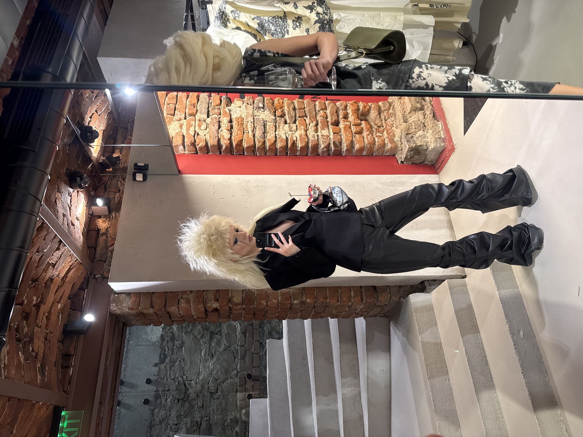 A female-presenting individual is depicted in a full-length mirror selfie, positioned slightly to the left of the frame. She has voluminous, spiky blonde hair, dramatic dark eye makeup with dark streaks extending below her eyes, and is wearing a black, open blazer revealing her décolletage, paired with baggy, ruched black pleather trousers that pool at the ankles over dark footwear. Her right hand holds a black smartphone, positioned to capture the selfie, while her left hand holds a small silver bowl containing red items, possibly berries, and also secures a small black handbag.
The environment combines historical and modern industrial elements. To the left of the individual, a concrete staircase with several visible steps descends towards the foreground. Behind the staircase, a rough-hewn stone wall is visible. Directly behind the individual, a textured brick wall with exposed mortar rises, intersected by a narrow, vertical red panel on the right. The ceiling features exposed brick arches, dark steel beams, and black industrial ventilation ducts, with multiple modern spotlights illuminating the space. A green illuminated sign, partially visible, displays "KIT" on an overhead beam.
In the mirror's reflection on the right side of the image, the individual's side profile, appearing as a second person, is visible. This reflection shows a person holding a clear stemmed glass, possibly a wine glass, and a sage green shoulder bag. The reflected person wears a sleeveless top with a black and white abstract or tie-dye pattern. Behind this reflection, a clothing rack with various garments, including white, cream, and patterned pieces, is visible. The reflection also shows more of the brick wall, modern white wall sections, and two black CCTV cameras mounted on the ceiling.
Based on the architectural fusion of ancient stone walls, exposed brick arches, modern concrete stairs, steel beams, and industrial design elements, the scene is located within a renovated historical building in central Sofia, Bulgaria. The presence of clothing racks suggests it is likely a high-end fashion boutique, a concept store, or an exhibition space integrating historical layers, commonly found in areas such as near the Ancient Serdika Complex.