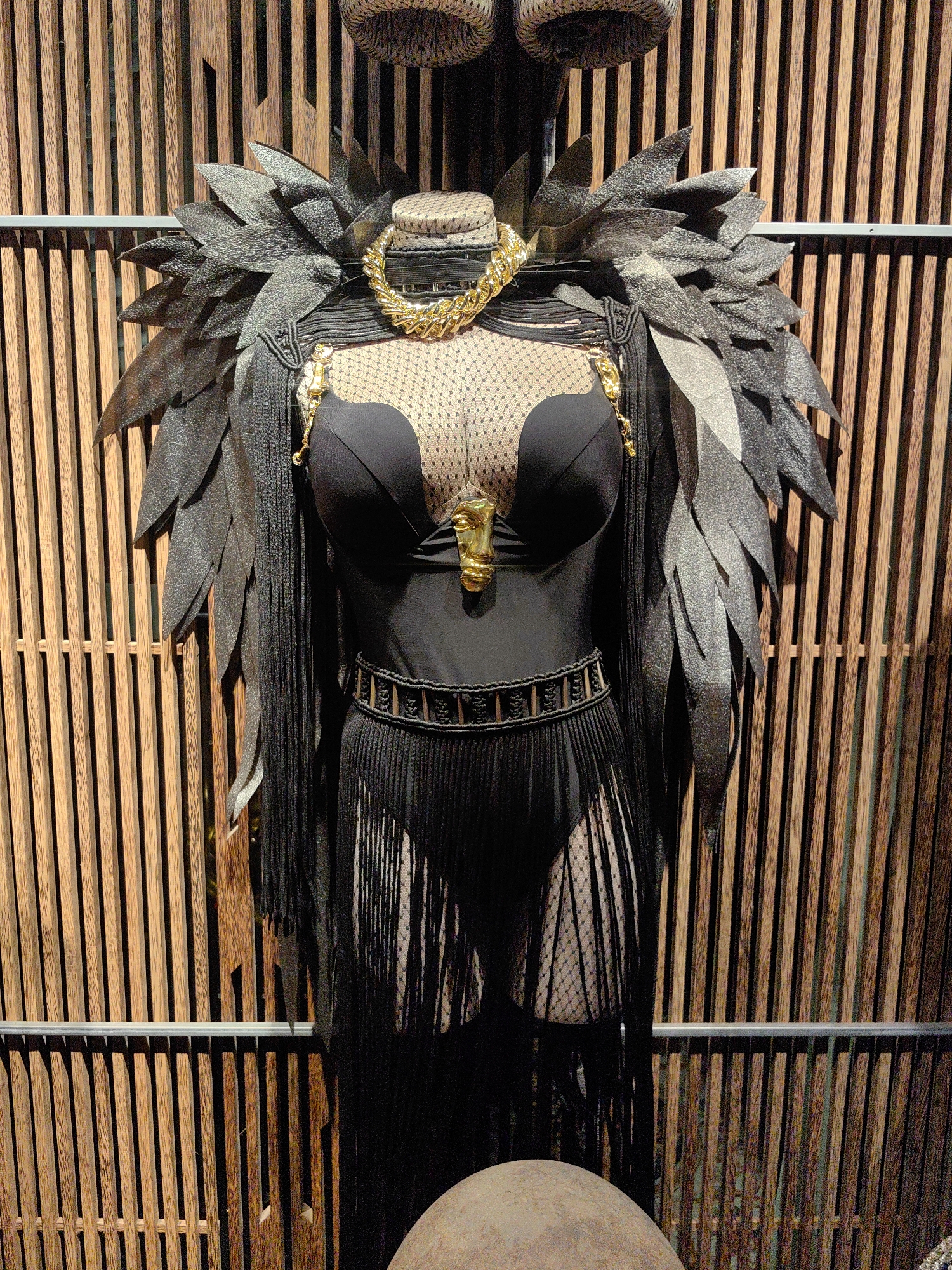 This media file shows a mannequin dressed in a dramatic black costume, likely for a performance or display, in Baan Mae Nam, Thailand. The costume features a black, strapless one-piece garment with a plunging neckline, adorned with a delicate black mesh overlay. A large, ornate gold chain necklace encircles the mannequin's neck. Extending from the shoulders are large, layered black feather-like accents, giving the impression of wings.
Below the chest, the outfit is cinched with a wide, intricately woven black belt. A prominent gold pendant shaped like a face is attached to the bodice. The garment continues into a skirt or shorts-like bottom made of sheer black fabric with a polka dot pattern, overlaid with numerous long, black fringe strands that reach down towards the bottom of the frame. The overall aesthetic is theatrical and bold.
The mannequin is positioned in front of a backdrop of vertical wooden slats, creating a textured, naturalistic setting. A thin metal bar runs horizontally across the wooden slats. The lighting suggests an indoor environment, possibly a shop or exhibition space. No text is visible in the image. The time of day and weather are not discernible. The focus is on the elaborate costume and the mannequin presenting it.