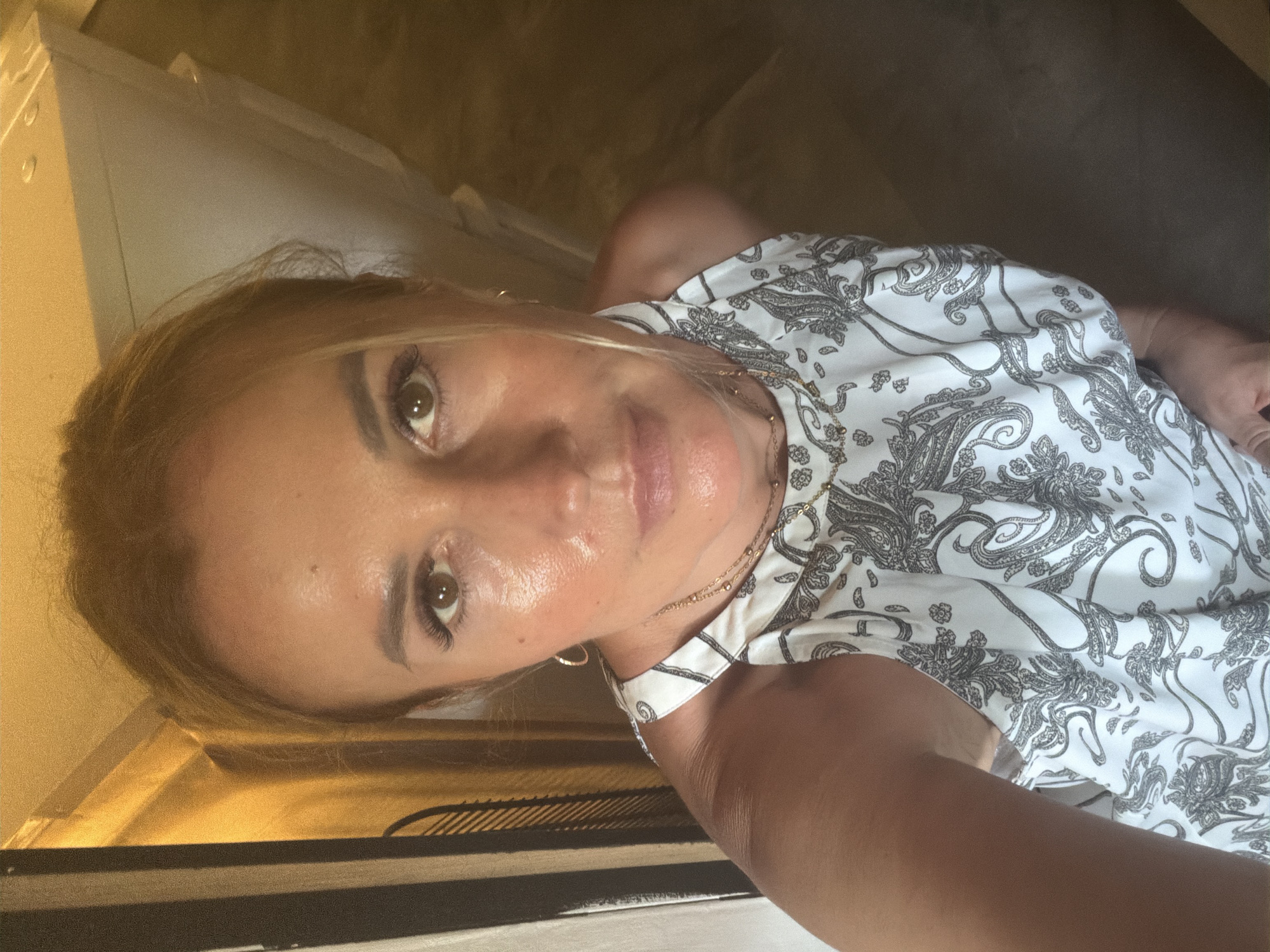 This media file is a selfie taken by a woman with blonde hair, who appears to be in an indoor setting. She is wearing a white top with a black paisley print and a delicate gold necklace. Her makeup includes mascara and lipstick. The lighting suggests it might be daytime, possibly with artificial light contributing to the warm glow. The background is blurred, but hints of furniture and what looks like a textured surface are visible, creating a somewhat intimate and casual atmosphere. The woman is looking directly at the camera with a neutral expression. The image is oriented sideways.