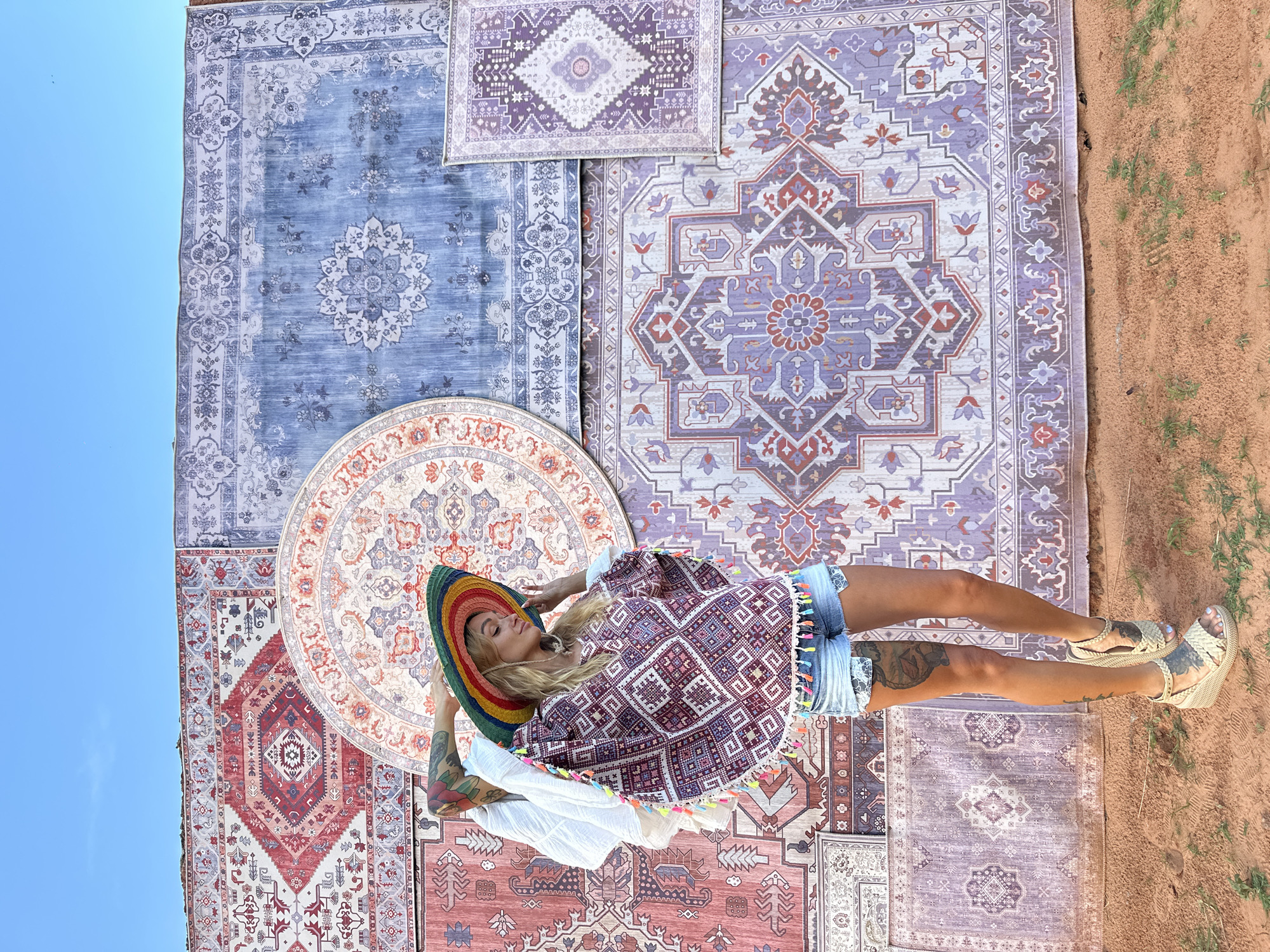 A woman stands in front of a wall covered in several large, patterned rugs in Mui Ne, Vietnam. The woman, with blonde hair and tattoos on her arms and legs, is wearing a colorful, wide-brimmed hat, a patterned poncho with colorful tassels, and denim shorts. She is wearing wedge sandals and has her hand raised to her hat. The rugs on the wall feature intricate, traditional patterns in various colors, including red, blue, and purple. The ground in front of the wall is sandy and has some sparse green vegetation. The sky above is a clear blue.