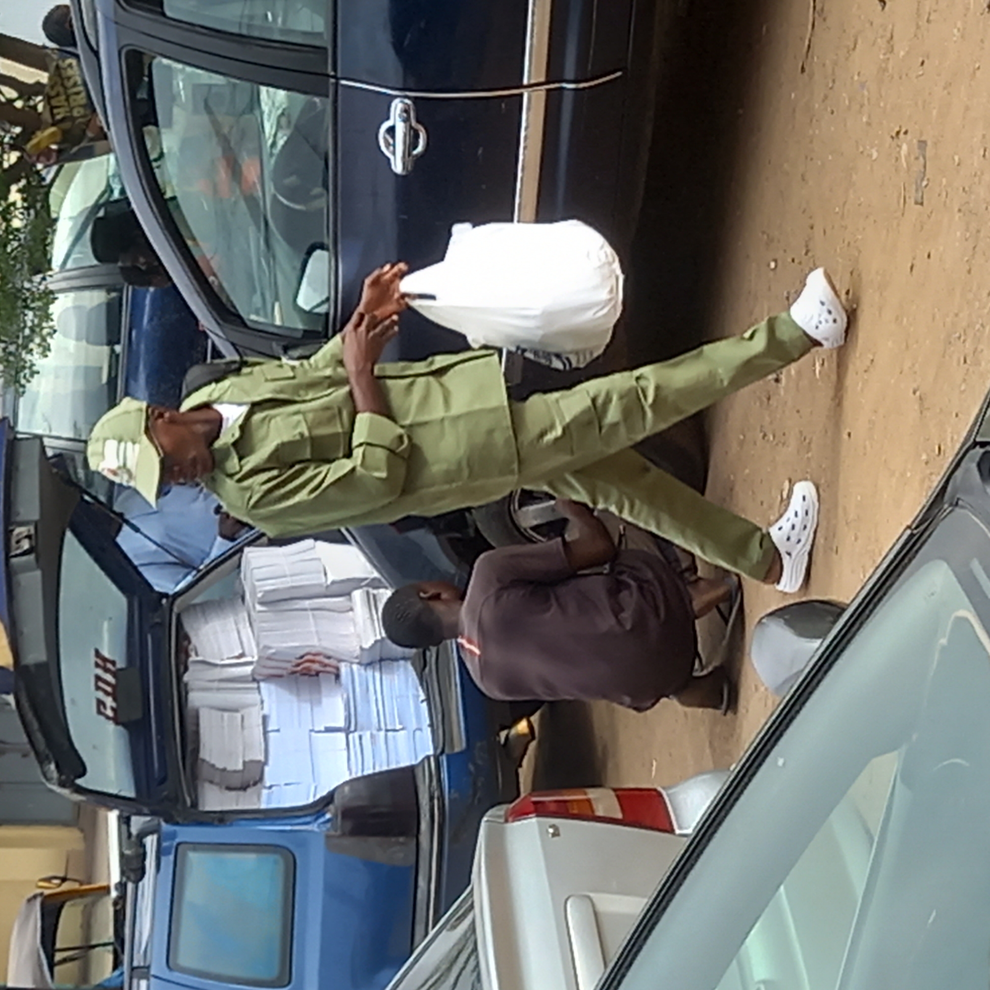 In Dadi Riba, Nigeria, a man in a green uniform, possibly associated with the National Youth Service Corps (NYSC), is holding a white bag beside a dark blue car. Another individual is kneeling near the car, while stacks of paper are visible inside the vehicle. A white car is partially visible on the right side of the image. The setting appears to be outdoors, possibly a street or parking area.