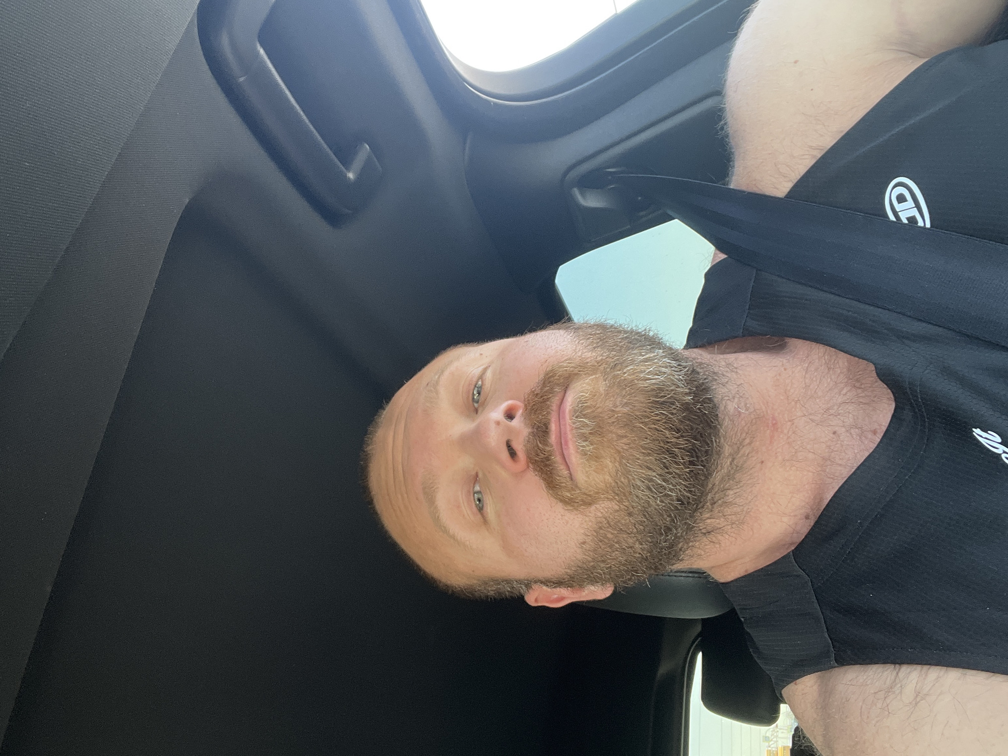A man with a beard is wearing a black t-shirt and a seatbelt in a car. The car has a sunroof. The location is not identifiable from the image.