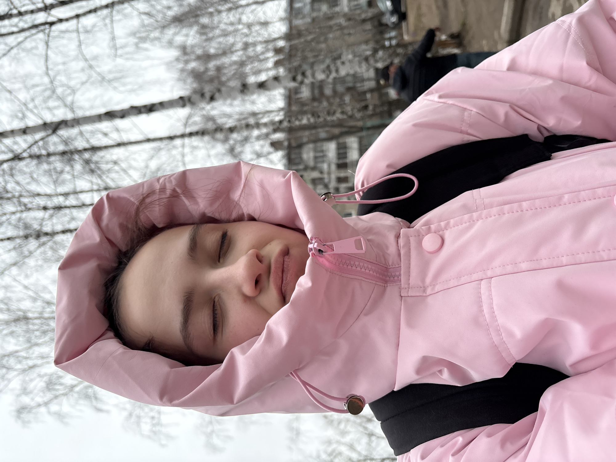 The image captures a close-up of a young individual resting outdoors, with their eyes closed and a peaceful expression. They are enveloped in the hood of a vibrant pink, padded jacket. Notable details on the jacket include a pink plastic zipper, pink drawstrings with metallic toggles, and a pink snap button. A black garment is visible beneath the opening of the jacket.
The background is softly blurred, depicting a wintry or early spring scene with numerous bare tree branches against a light, overcast sky. In the distant background, indistinct buildings and a blurred figure in dark clothing are faintly visible, suggesting an urban or suburban environment. The overall impression is of a quiet moment of repose in a cool, outdoor setting, contextualized as Chaikovsky, Russia. There is no visible text.