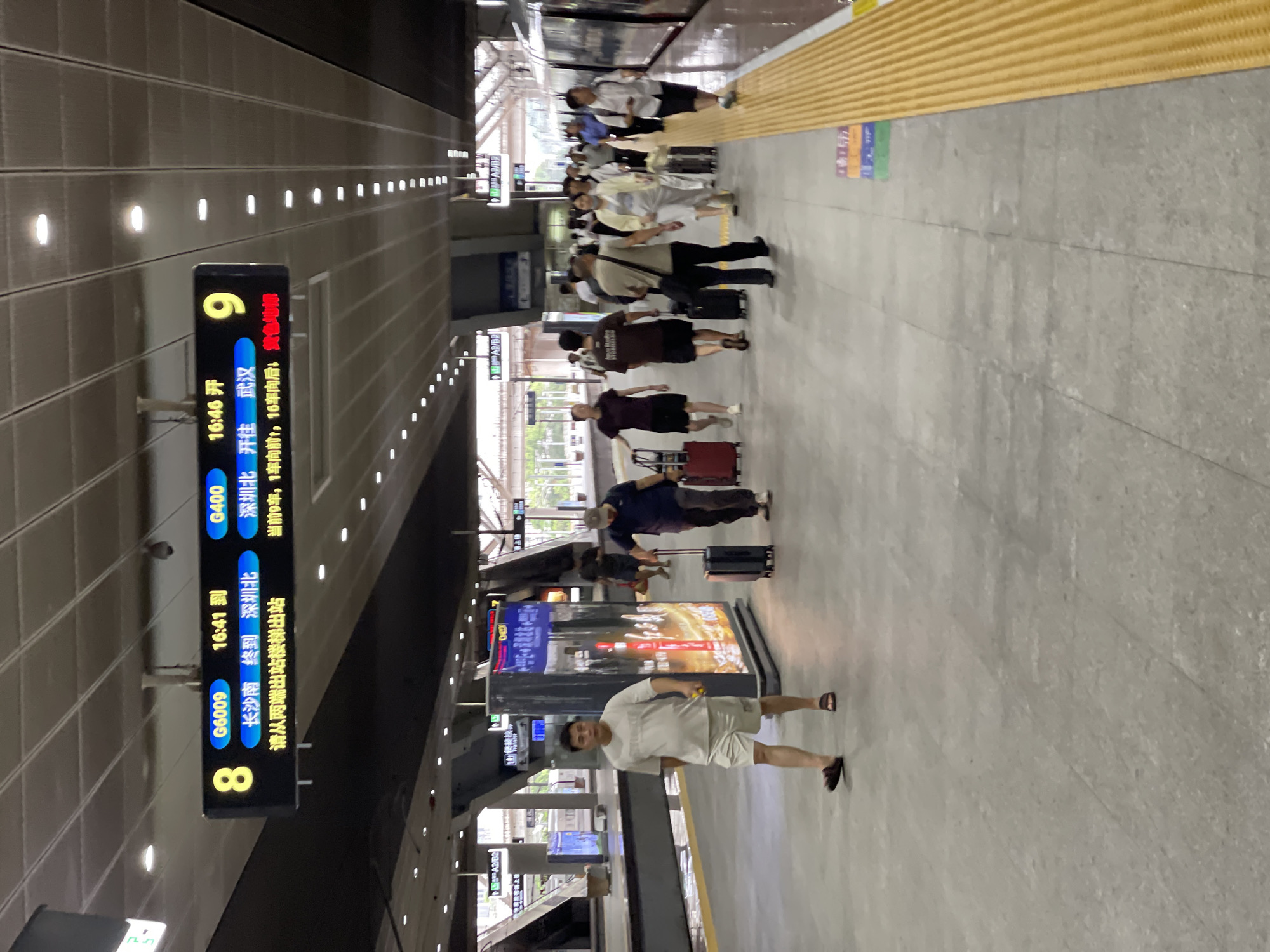 The image depicts a bustling train station platform in Longhua District, China. Several individuals, mostly dressed in casual attire, are waiting or walking along the platform. Some are seen with luggage, suggesting they are travelers. The scene appears to be indoors, under a covered structure, with artificial lighting from above. A yellow tactile paving runs along the edge of the platform, a common safety feature in public transportation areas.
A prominent digital display board shows train information. It indicates that train G6009, arriving at 16:41 from Changsha South, will terminate at Shenzhen North. Another train, G400, is scheduled to depart for Wuhan at 16:46 from Shenzhen North. The time of day is late afternoon, as indicated by the train schedules. The overall atmosphere seems calm but busy, with people engaged in their travel plans. The ground is paved with concrete tiles. The lighting suggests it could be daytime, but the covered nature of the station makes it difficult to ascertain the exact weather conditions. The presence of multiple train lines and the efficient display of information point to a well-developed public transport hub. The text on the signs is in Chinese, providing essential details for passengers.