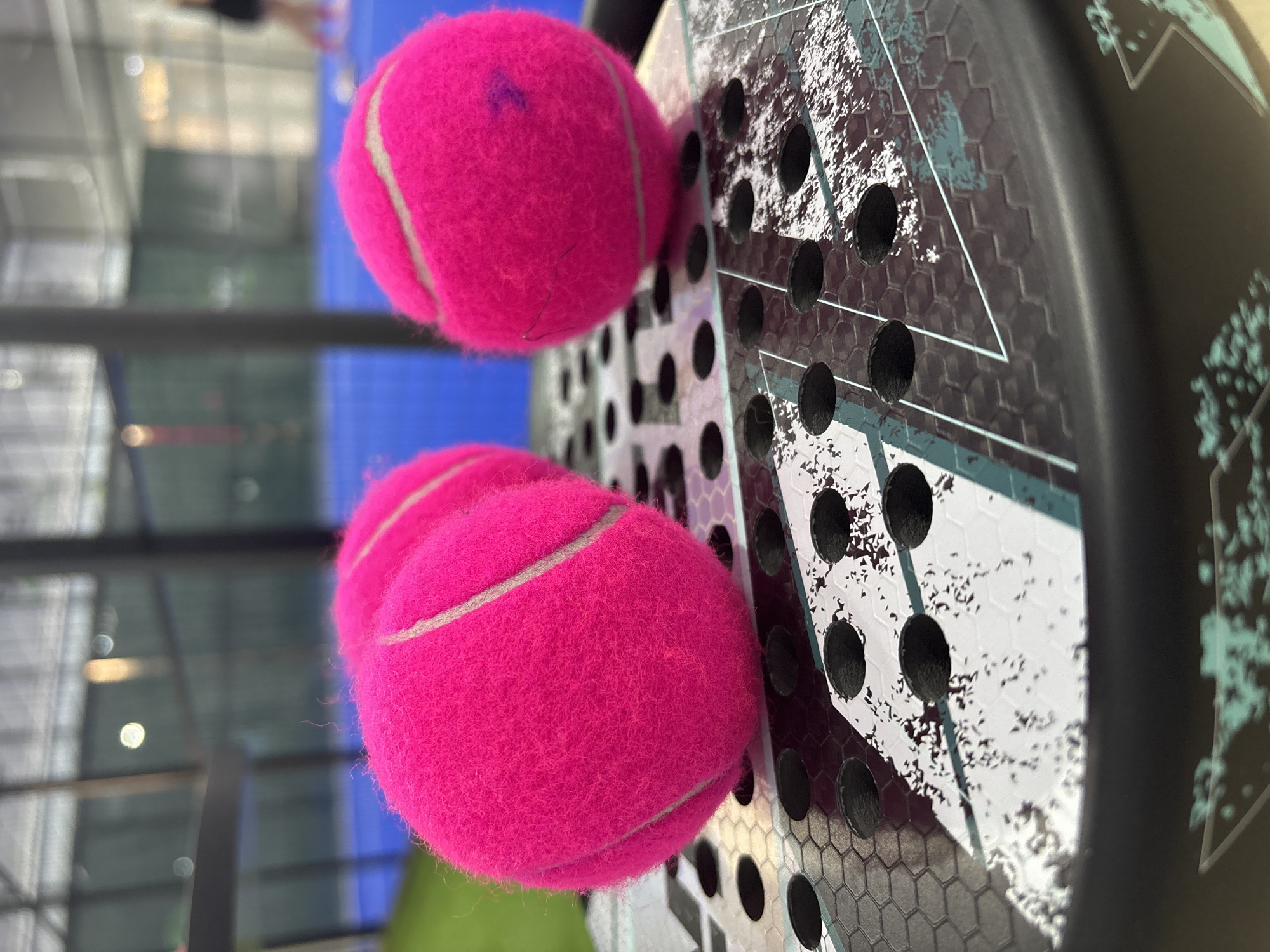 The image displays a close-up, angled view of a black padel racket with a patterned surface featuring hexagonal shapes and abstract white and teal designs. Three bright pink padel balls are positioned on the strings of the racket. One ball is in the foreground, with two others slightly behind and to the left. The balls appear fuzzy and have a visible white seam. A faint purple mark is present on the uppermost ball.
The background is out of focus, but suggests an indoor sports environment. A blue court surface is visible, along with structural elements of what appears to be a building, possibly with large windows or skylights, suggesting daytime. The overall impression is of a sports setting, likely a padel court. There is no visible text in the image.