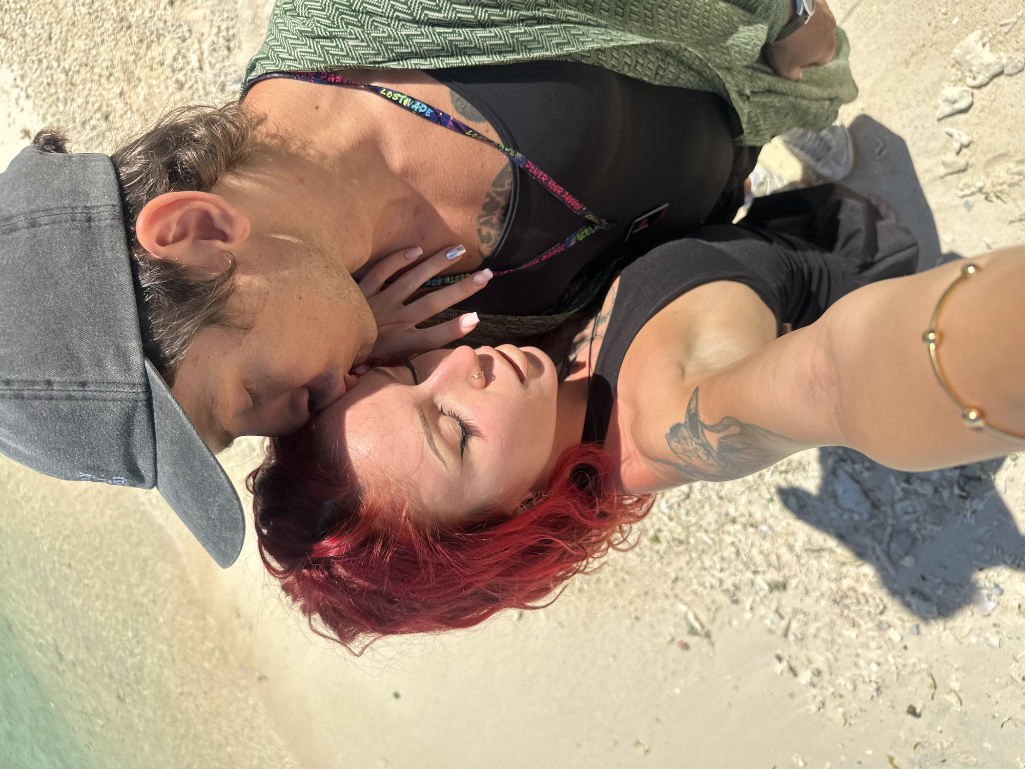 A couple is lying on a sandy beach. The woman has red hair and a black tank top. The man is wearing a grey baseball cap and a green patterned shirt. They are both lying on their backs and are in close proximity to one another. The background is a sandy beach with some visible rocks and light green water. The location appears to be in Unknown, Indonesia.