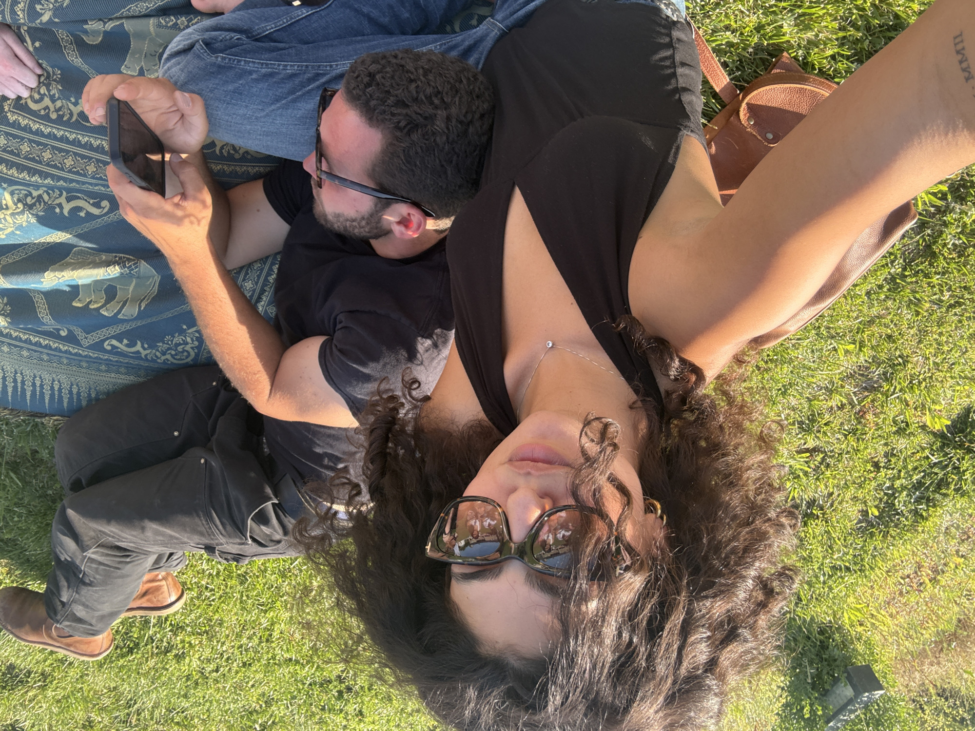 A medium overhead outdoor shot depicts a man and woman lying on a blanket, likely in a park in Los Angeles, USA. The man, wearing black framed glasses, is lying down and looking at a smartphone held by another person in the frame. He is wearing a black t-shirt and has a beard. The woman, presumably taking the photograph, has long, dark curly hair and is wearing sunglasses. She is wearing a black top with a delicate necklace visible at her neckline. Both are on top of a patterned blanket with elephant designs. The woman has her arm stretched out to take the photo, and a brown leather satchel is visible in the background. The scene is illuminated by bright sunlight, casting shadows on the grassy area.