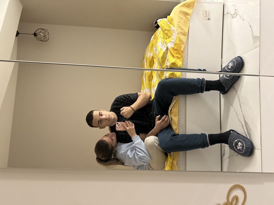 A mirror selfie captures two individuals in a modern bedroom, described as being located in Sofia, Bulgaria. A male, with short dark hair, sits on the edge of a white bed frame, facing forward. He is dressed in a dark, patterned short-sleeved t-shirt, dark blue jeans with cuffed hems, black socks, and dark blue slippers featuring a white circular logo that reads "NEW YORK CREW SINCE 1978." His left hand is clenched into a fist, and he wears a red bracelet on his right wrist. A female is positioned behind and to the left of the male, resting her chin on his left shoulder. She has light brown hair pulled into a ponytail and is wearing a light blue long-sleeved sweatshirt and light-colored pants. She holds a light pink smartphone, which is used to take the reflection photograph, positioned in front of the male's chest. The bed is covered with a bright yellow duvet patterned with white floral designs. The room's environment includes neutral-toned walls, a floor covered in large, white marble-patterned tiles, and a black geometric wireframe pendant light fixture suspended from the ceiling in the upper right portion of the reflection. A decorative gold-colored accent is partially visible on the left edge of the mirror's frame.