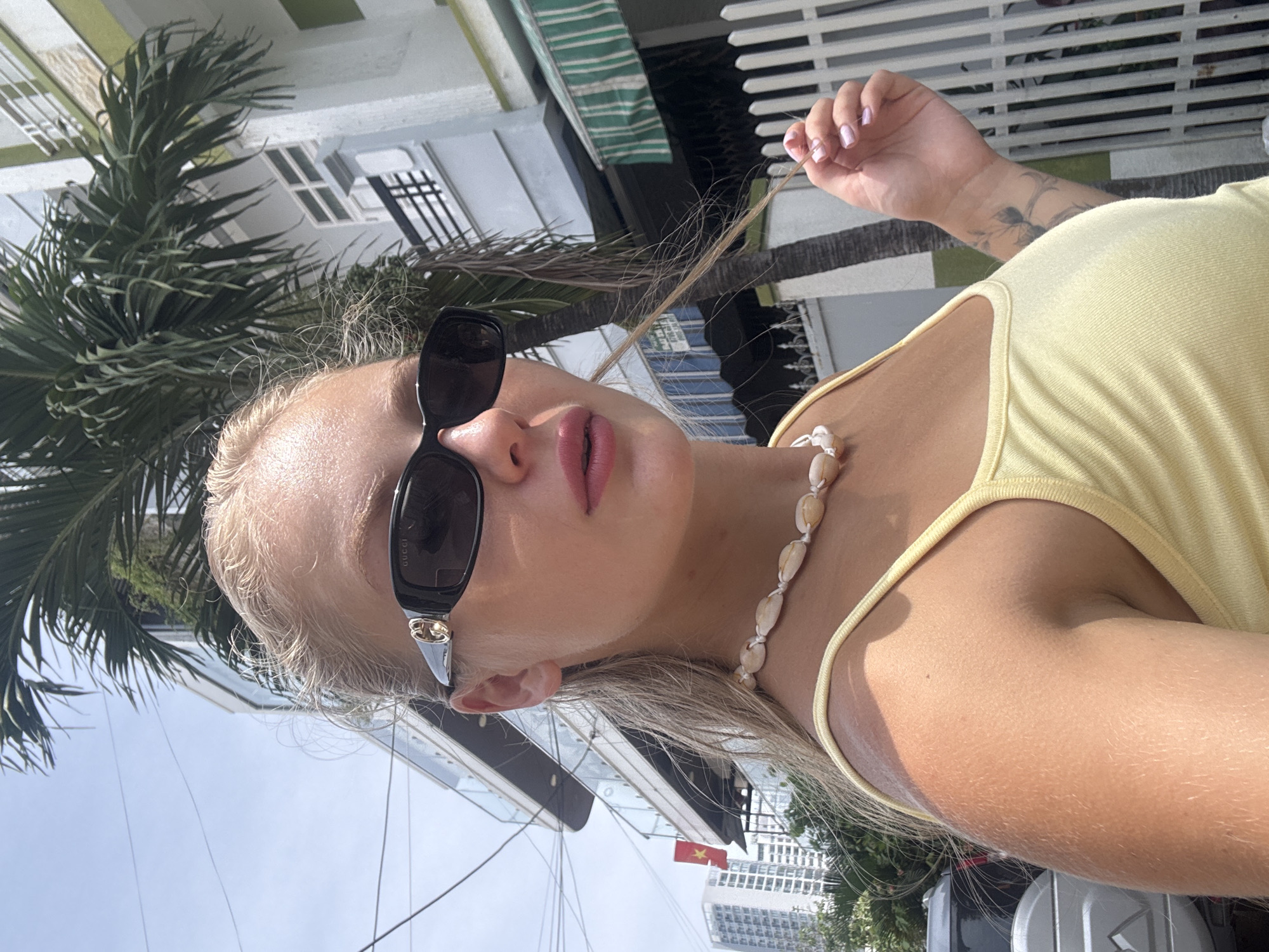In a selfie taken outdoors, a young Caucasian woman with blonde hair tied back and wearing a yellow tank top and sunglasses poses with a slightly blurred background. She is adorned with a seashell necklace. Her left arm, with a tattoo of a floral design, is visible. She is holding a strand of her hair with her right hand. The environment suggests a city setting, with buildings and a palm tree visible. A glimpse of a red flag with a yellow star, possibly the Vietnamese flag, is seen on a building in the distance, hinting at the location being in Vietnam. The image is captured on a sunny day, with the bright sunlight illuminating the scene.