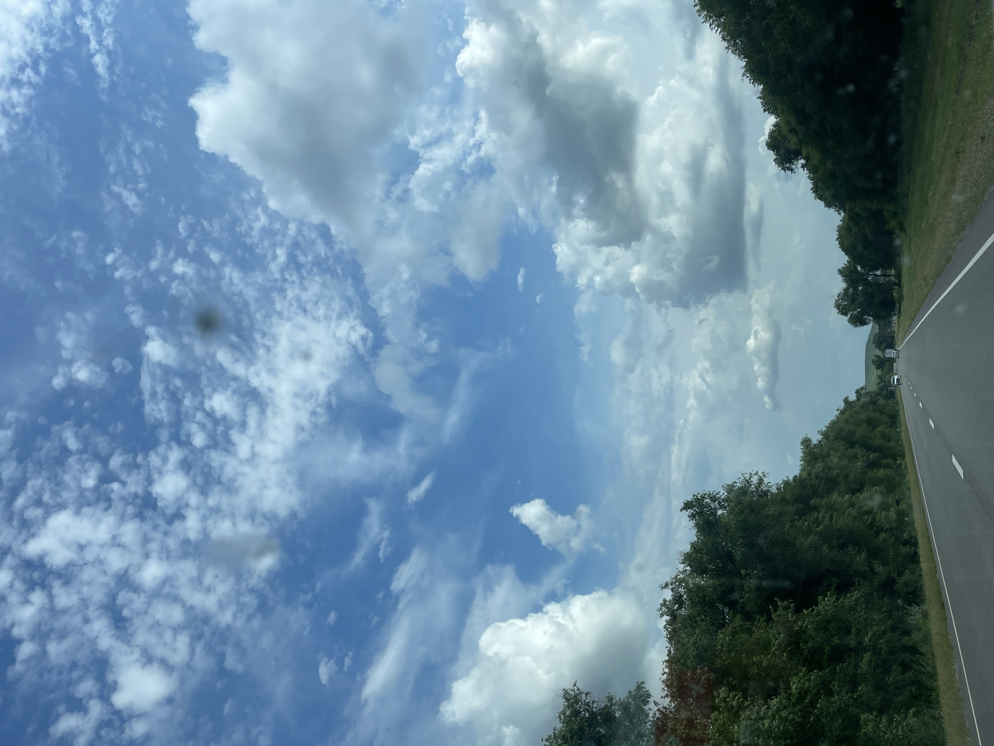 The image depicts a scenic view from a vehicle traveling along a road in Donskoe, Russia. The perspective is from inside the car, looking upwards and to the right. The sky is a dominant feature, filled with a mix of fluffy white cumulus clouds and thinner, wispy cirrus clouds against a vibrant blue backdrop. Sunlight filters through, suggesting it is daytime, likely midday or early afternoon.
The road itself is visible on the right side of the frame, an asphalt surface with white lane markings. It appears to be a relatively straight stretch, bordered by a strip of green grass and a dense line of dark green trees. These trees form a thick canopy, creating a natural barrier along the roadside. The foliage suggests a warm season, possibly summer.
In the distance, along the road, a few vehicles can be faintly seen, indicating ongoing travel. The overall atmosphere is one of a peaceful journey through a verdant landscape under a dynamic sky. There is no visible text in the image. The scene evokes a sense of calm and the simple beauty of nature.