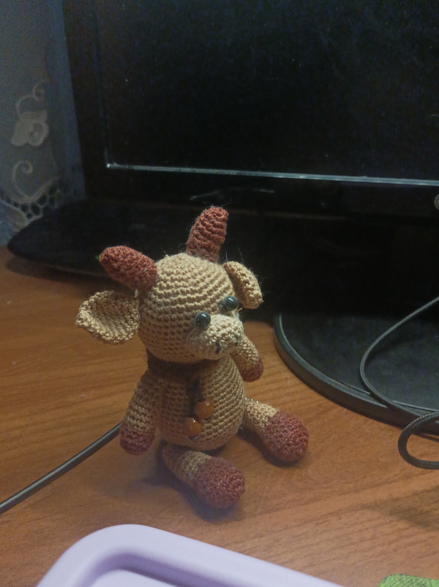 A small, handmade crocheted stuffed animal is centrally featured, sitting on a wooden surface. The animal has a light beige body, head, and one ear, contrasted with reddish-brown limbs, two small horns, and the other ear. It has small, dark, bead-like eyes and wears a simple brown collar with two amber-colored beads on its chest.
In the background, a large, dark electronic screen, likely a computer monitor, occupies the upper right portion of the frame. Its dark, circular base is visible on the right side of the wooden surface, with a black cable running across the desk towards it. To the upper left, a sheer, patterned curtain or piece of fabric is partially visible against a dark background.
A portion of a light purple plastic container is visible in the bottom-left corner of the image. The setting is indoors, on what appears to be a desk or table, with ambient or artificial lighting creating a somewhat subdued atmosphere. There is no activity taking place, as the stuffed animal is stationary. No specific geographical cues are evident beyond a typical indoor environment. No text is visible in the image.