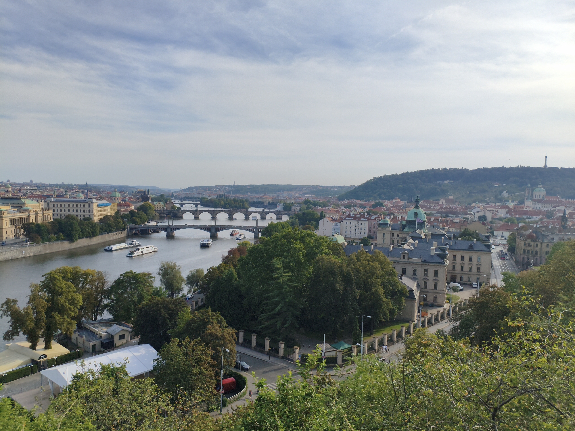 This is an elevated, wide-angle, daytime, eye-level shot of a city landscape in Prague, Czechia, overlooking the Vltava River and multiple bridges. The sky is a pale blue with scattered white clouds.
In the foreground, lush green trees and foliage dominate, with some appearing as if viewed from a hillside. To the left and center-left of the foreground, several white and tan tents are set up, suggesting an outdoor event or marketplace. A paved pathway with bollards runs alongside the tents.
Dominating the midground is the wide, calm Vltava River, with several tour boats and barges moving on its surface. Several bridges span the river, the closest being a wide, stone bridge with multiple arches. Further in the distance, more bridges are visible, including one with a distinct railing design.
On the far bank of the river, the cityscape unfolds. Numerous buildings with red-tiled roofs and light-colored facades line the river and extend into the background. Architectural details include domed roofs and classical building designs. One prominent building in the mid-right midground features a dark gray roof and beige walls with numerous windows. Further to the right and in the distance, a tall, slender tower-like structure is visible on a distant, tree-covered hill, which is likely the Petřín Lookout Tower. The overall impression is a bustling European city on a pleasant day.
