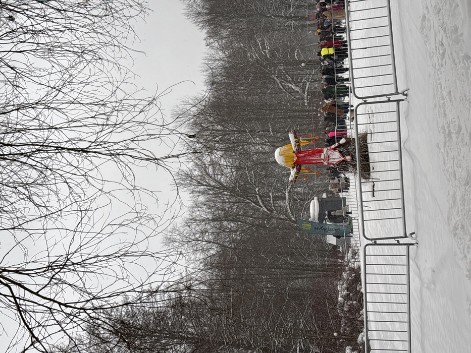 A winter scene shows a crowd behind a metal fence. A large straw figure with a white head, red and gold dress, and orange arms stands prominently. Bare trees fill the background. Snow covers the ground. A sign with Cyrillic text is visible.