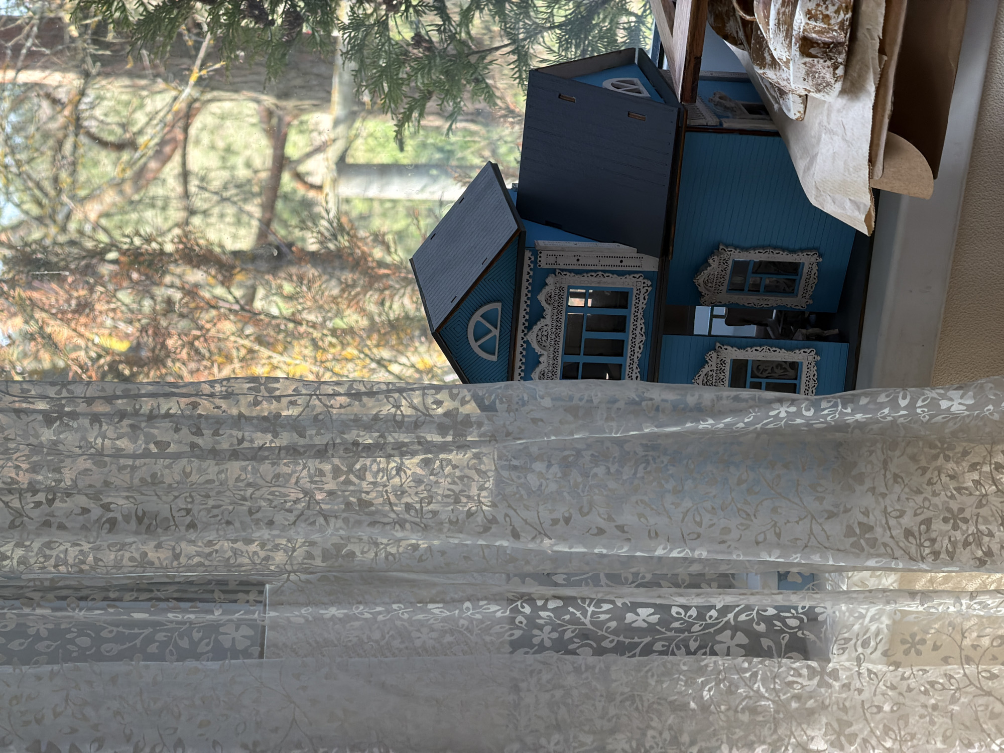 The image captures an indoor scene looking out a window. In the foreground, a translucent white sheer curtain, possibly a Roman blind, features a delicate, repeating floral and leaf pattern. This curtain covers the lower portion of the window, softly filtering the light.
Above the curtain and positioned near the right side of the window is a multi-part blue model house or dollhouse. The house is constructed from wood, painted a vibrant blue, and adorned with intricate white decorative trim, particularly around the windows and gables, giving it a charming, ornate appearance. Some sections of the model house show exposed wooden edges, suggesting it might be a kit, incomplete, or disassembled. Nearby, on the far right, crumpled brown paper or packaging material is visible.
Through the window, beyond the model house, an outdoor scene unfolds. Bare tree branches with some visible green foliage (possibly juniper or cypress) indicate a natural setting, likely in late autumn or early spring. The distant background shows a blurred landscape of more trees and greenery. The overall lighting suggests it is daytime, with a soft, diffused light illuminating the scene. There are no people present, and no specific activity is taking place; it's a static composition focusing on the interplay between the indoor objects and the outdoor view. No visible text is present in the image.