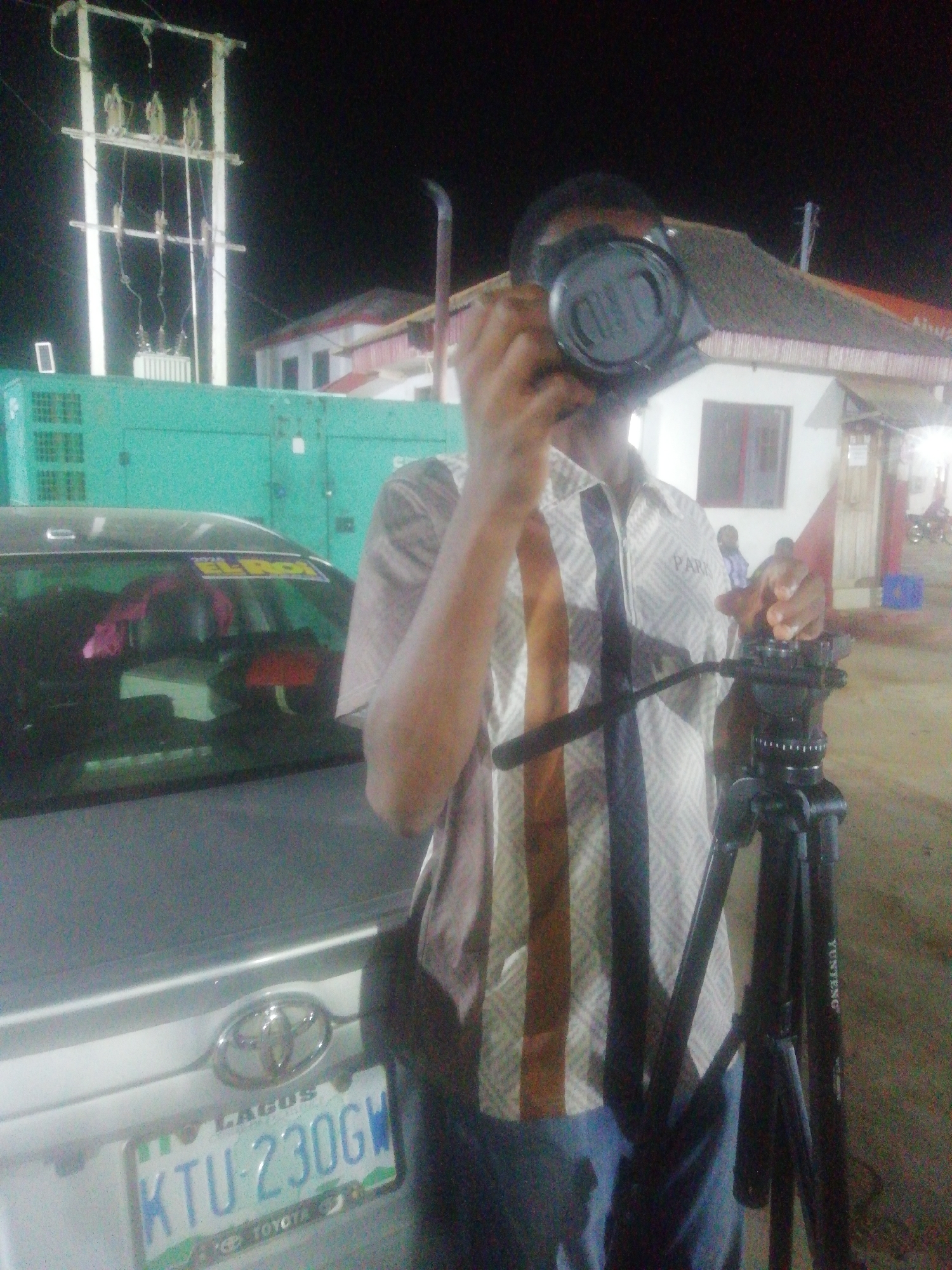 A man stands in front of a light silver Toyota car with a Lagos, Nigeria, license plate number KTU-230GW. He is holding a black camera up to his face with his left hand and adjusting a black tripod with his right hand. The man is wearing a short-sleeved shirt. Behind the car is a green generator, an electrical transformer, and buildings. The tripod has the word "Yunteng" written on one of the legs. The man appears to be setting up the camera to record footage. The location is identified as Ikorodu, Nigeria, based on the Lagos license plate. The sky is dark, suggesting it is nighttime.