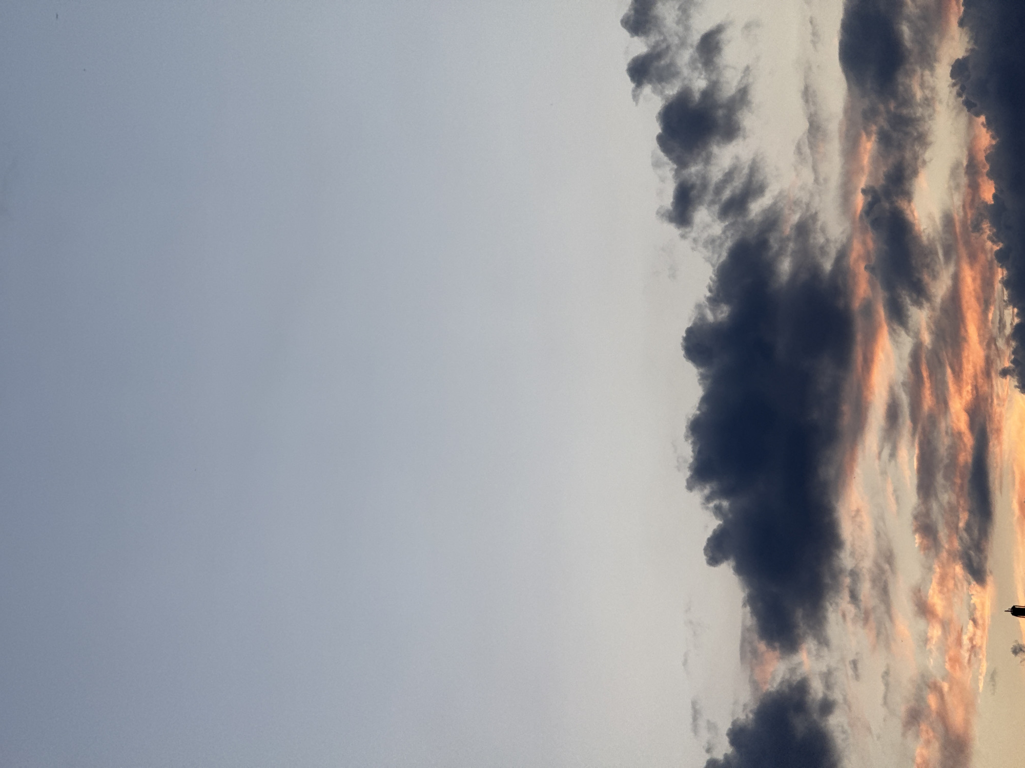 A cloudy sky with a touch of sunset colors is visible. The bottom portion of the image shows dark grey clouds interspersed with streaks of orange and pink, indicating the presence of sunlight filtering through. A very small portion of a tall, dark structure, possibly a tower or antenna, is visible at the very bottom left. The upper portion of the image is a clear, pale blue sky. The scene appears to be an outdoor, wide-angle shot taken from ground level looking upwards.