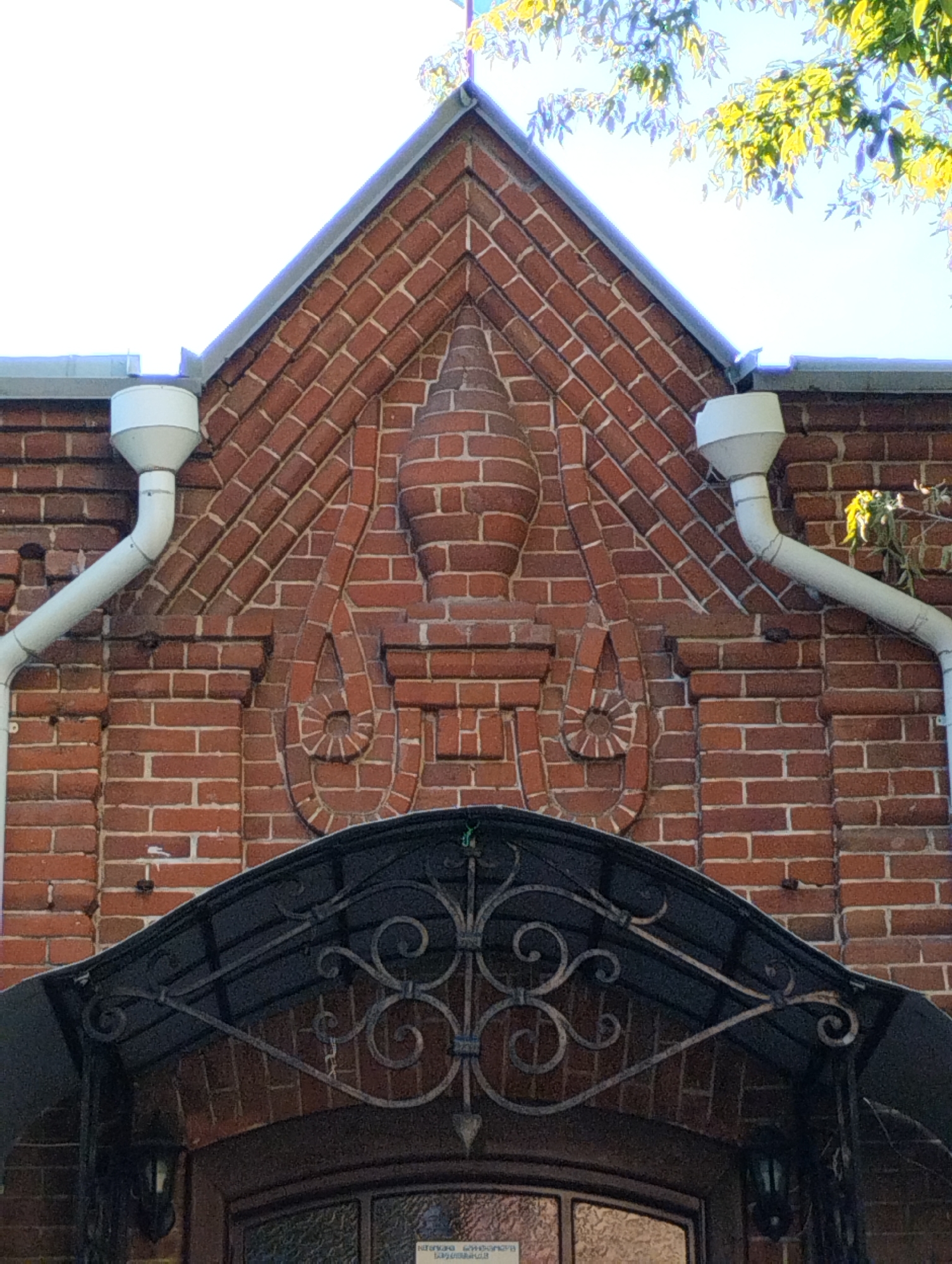 The image shows the upper facade of a brick building, likely a historical or architecturally significant structure, located in Kostanay, Kazakhstan. The building features ornate brickwork, with diagonal patterns forming a triangular pediment. Centrally positioned within the pediment is a decorative brick element resembling a stylized urn or finial. Flanking this central element are two downward-curving decorative brick motifs that end in spiral shapes. White downspouts are visible on either side of the facade, directing water from the roof. Below the pediment, a dark, wrought-iron awning with intricate scrollwork extends over an entrance. The awning is supported by decorative metal brackets. Two dark-colored outdoor light fixtures are visible hanging from the underside of the awning, one on each side. The entrance itself appears to be a door with a glass pane, partially visible at the bottom of the frame. The overall condition of the brickwork suggests age, with some minor imperfections visible. The presence of lush green foliage from a tree in the upper right corner indicates an outdoor setting, likely during daytime with clear sky or bright sunlight filtering through the leaves.