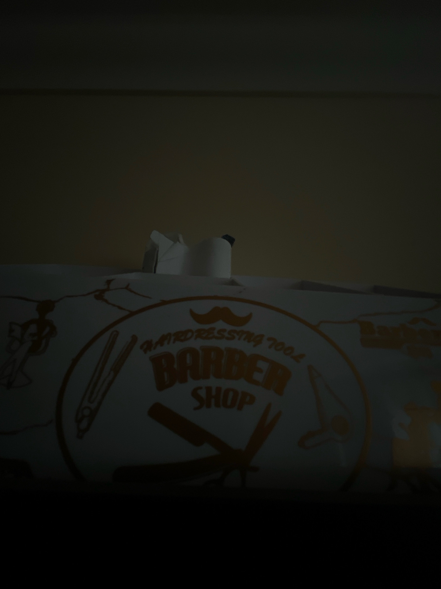 The image shows a roll of paper towels with a "Barber Shop" logo printed on it, set against a dark background. The logo features a mustache, scissors, and a comb. There are also some abstract figures printed on the paper towels. The scene is dimly lit, with the majority of the image being dark. There is no discernible urban environment or specific location within Balbriggan, Ireland. The image is a close-up of the product itself.