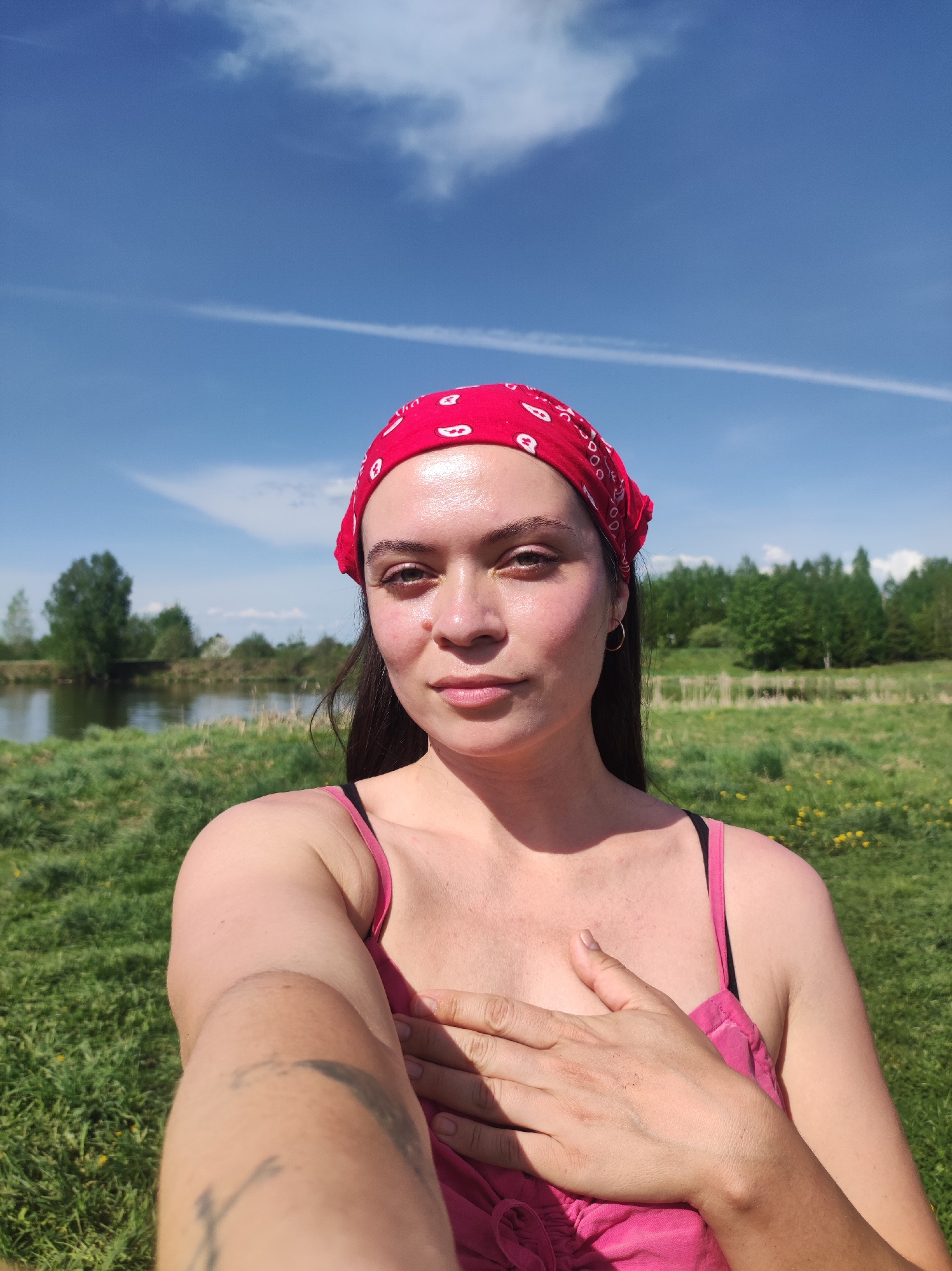 A young woman is taking a selfie outdoors. She has a red bandana on her head, dark hair, and is wearing a pink tank top. She is holding her hand to her chest. In the background, there is a grassy field, a body of water (possibly a lake or pond), and trees. The sky is blue with some clouds. The location is near Фоки, Russia, based on the natural environment.