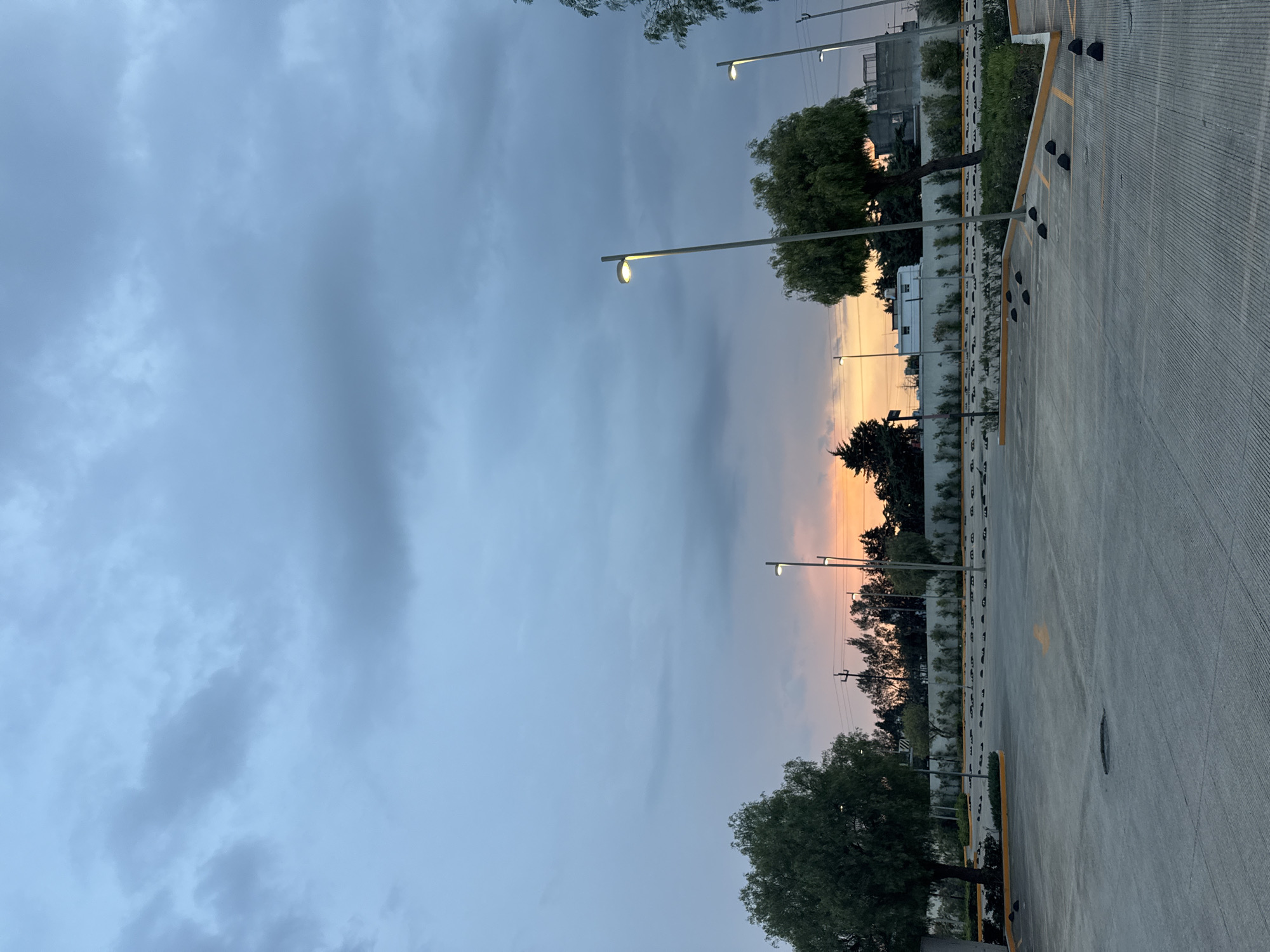 The image depicts a parking lot in Puebla, Mexico at dusk. The foreground is dominated by a concrete parking lot with yellow lines marking parking spaces and black parking bumpers in each slot. Metal light poles line the parking area. Beyond the parking lot are trees and vegetation silhouetted against the setting sun. The sky is overcast with gray and blue clouds.