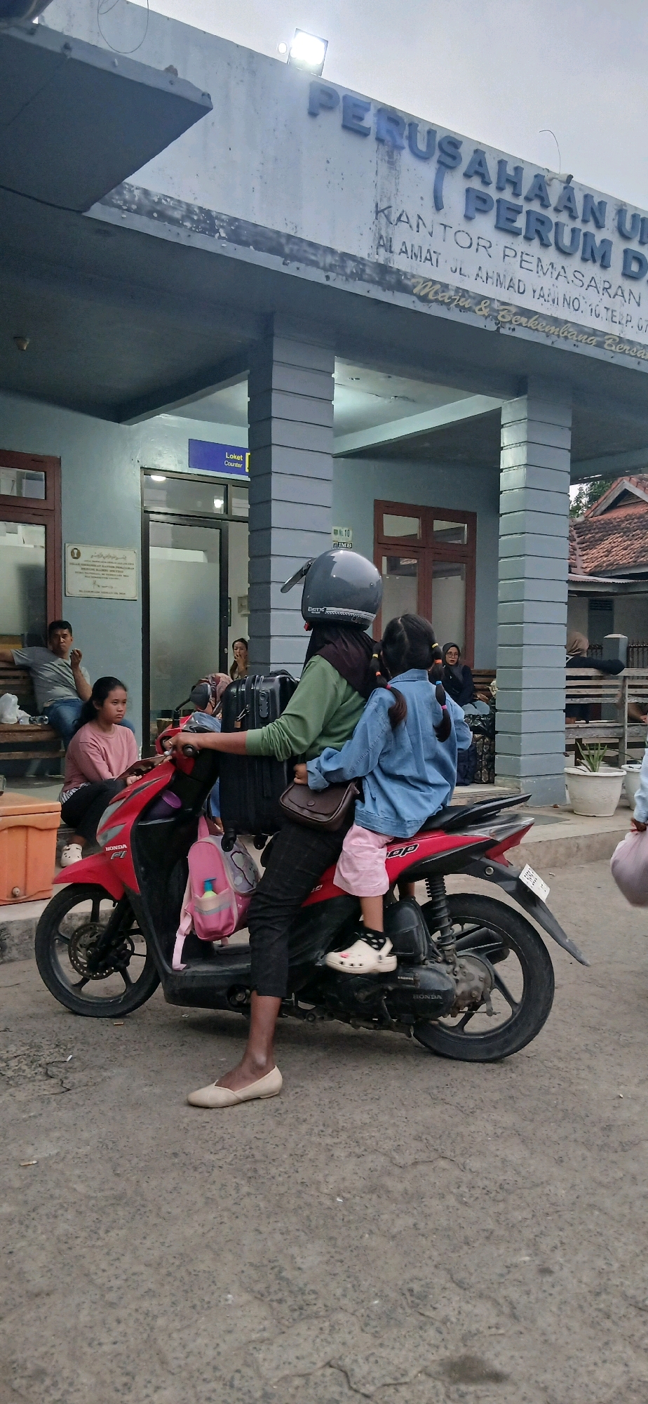 In front of a building with the text "PERUSAHAAN URUM" and "KANTOR PERUM DIN", a woman wearing a helmet and a green shirt, and a young girl wearing a blue denim jacket are sitting on a red motorcycle. The woman is holding the handlebars and the young girl is seated in front of her. The young girl has her arms around the woman. A black suitcase is attached to the back of the motorcycle. There are other people visible in the background sitting on benches and standing. The environment appears to be an outdoor waiting area. The building has columns and a sign that reads "Loket Counter". The ground is paved. This scene is located at the Kantor Perumahan Unit Perusahaan Umum Perum Perhutani, Jalan Ahmad Yani, No. 10, Tezep, Gunung Sugih, Central Lampung Regency, Lampung, Indonesia.