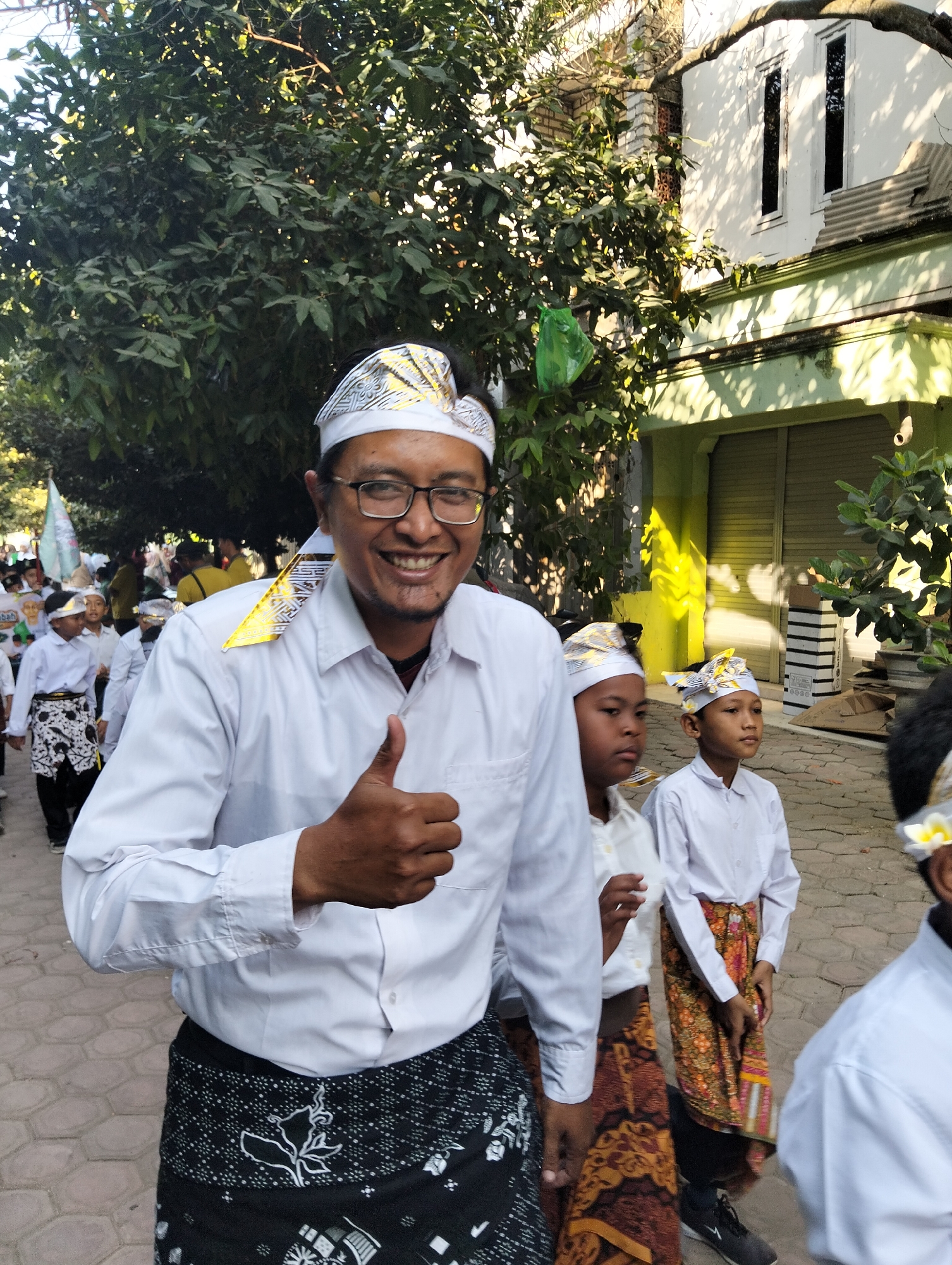 An adult male wearing a white long-sleeved shirt, glasses, and a white and gold patterned head covering gives a thumbs-up gesture while looking at the camera. He is wearing a black sarong with a white pattern around his waist. He is in the foreground of a procession of people, including several children, who are also wearing white shirts and patterned head coverings. The children are wearing sarongs of various colors and patterns. The procession appears to be walking on a paved walkway lined with trees and buildings. The buildings are painted in shades of white and green. The scene suggests a cultural or religious event taking place in Kranji, Indonesia.
