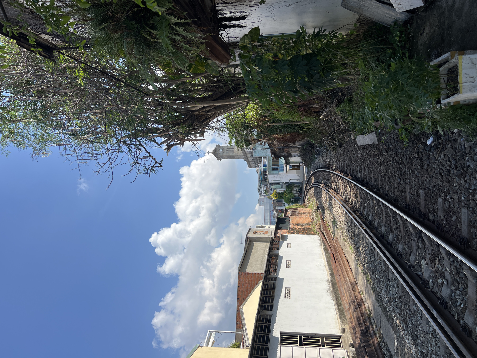 The image shows a railway track running through a densely built area. The sky is visible with scattered clouds. A church tower can be seen in the distance. The railway track curves to the right of the frame, bordered by buildings on one side and overgrown foliage on the other. The image is in the city of Nha Trang Ward, Vietnam.