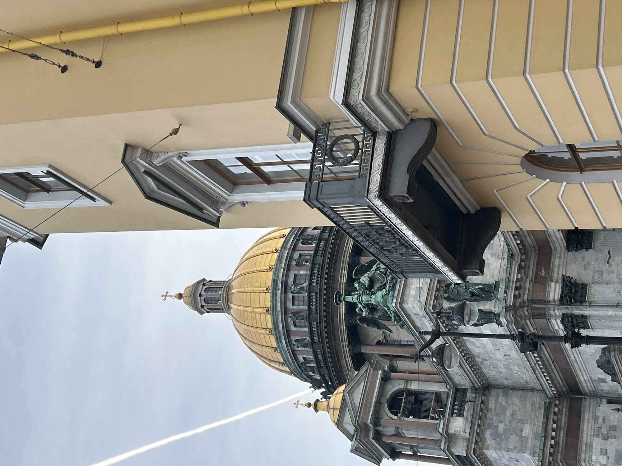 The image shows a view of a large, ornate building with a golden dome, likely a cathedral or a significant public building. The perspective is from below, looking upwards. The building appears to be made of stone and features intricate architectural details such as columns, statues, and decorative moldings. The golden dome is a prominent feature, topped with a cross.
In the upper part of the image, a light-colored building with a yellow facade is visible, featuring windows and decorative elements. There are also some overhead wires and fixtures attached to this building, suggesting it is a separate structure or an adjacent part of the same complex.
The sky is overcast, suggesting a cloudy day, and a white contrail from an aircraft is visible across the sky. The overall lighting is diffused, indicating it is likely daytime. There are no people clearly visible in the image. The scene appears to be set in an urban environment given the architecture of the buildings and the presence of overhead wires. The detailed stonework and golden dome suggest a location of historical or religious significance. The orientation of the image is such that the buildings are upside down relative to a typical viewing perspective.