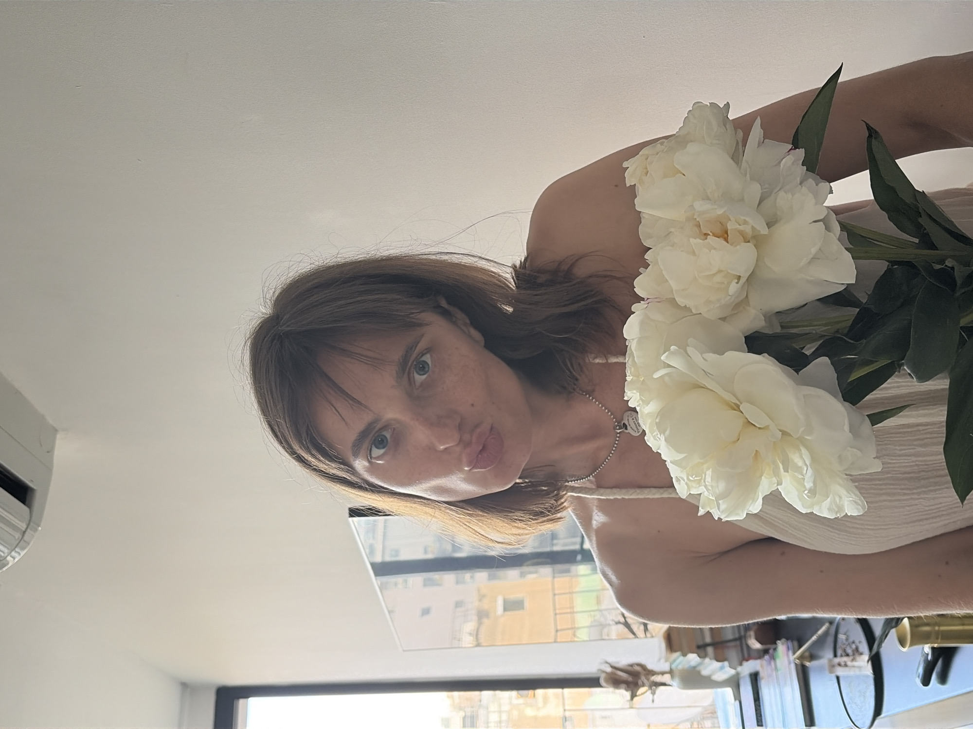 A woman with brown hair and blue eyes is holding a bouquet of white peonies. She is wearing a light-colored dress and a necklace with a heart-shaped pendant. She is puckering her lips and looking at the camera. The background is a wall and a window with a view of buildings. This appears to be an indoor shot, possibly in a residential apartment in Tel Aviv, Israel, given the architectural style visible through the window and the air conditioning unit on the ceiling.