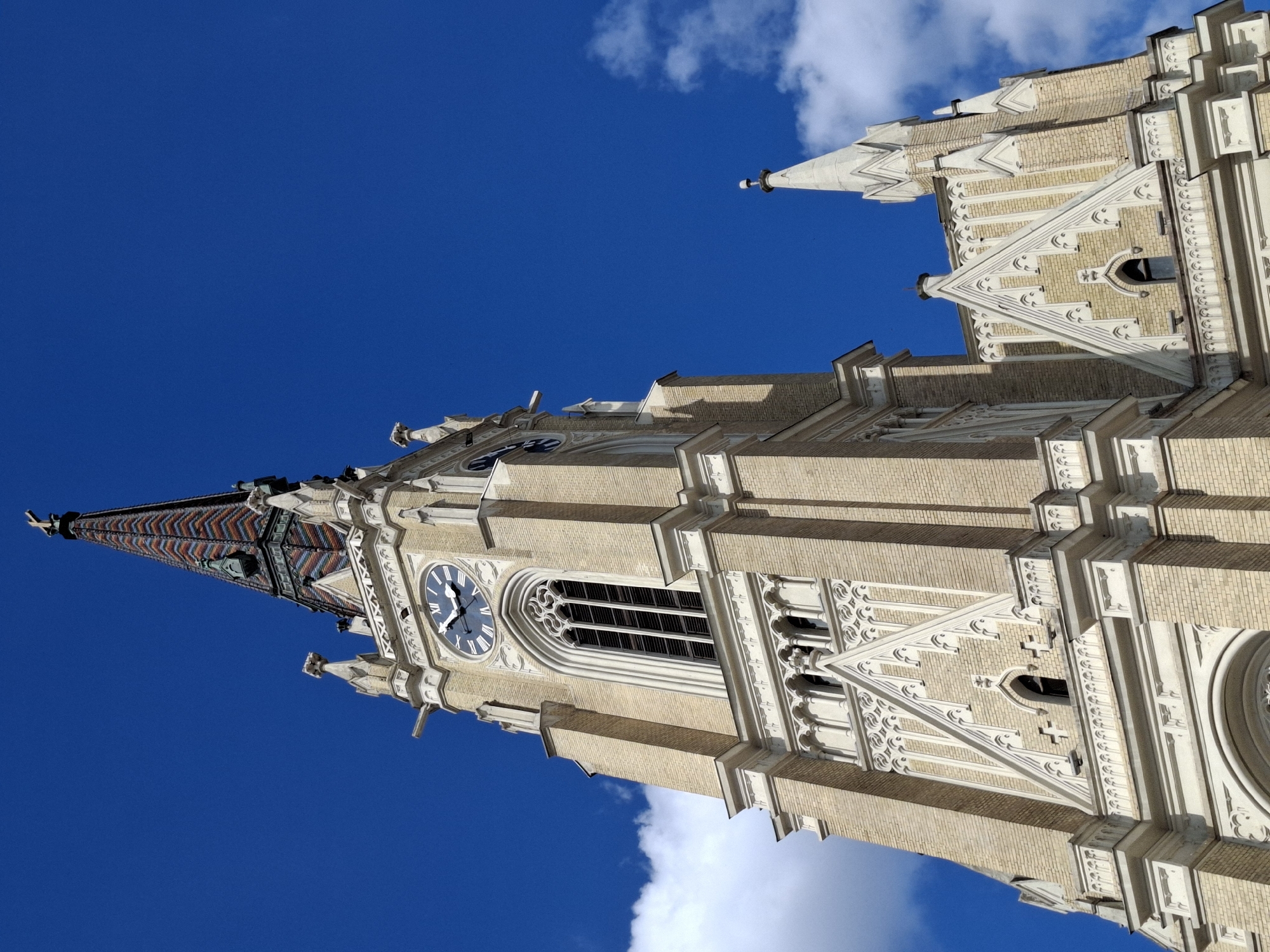 The image shows the architectural details of the Name of Mary Church in Novi Sad, Serbia. The facade is made of light-colored bricks and is adorned with intricate gothic-style carvings. A clock is visible on the tower, and the tall steeple is patterned with colorful tiles. The sky is bright blue with a few white clouds. The building is angled against the sky, giving a worm's eye view.