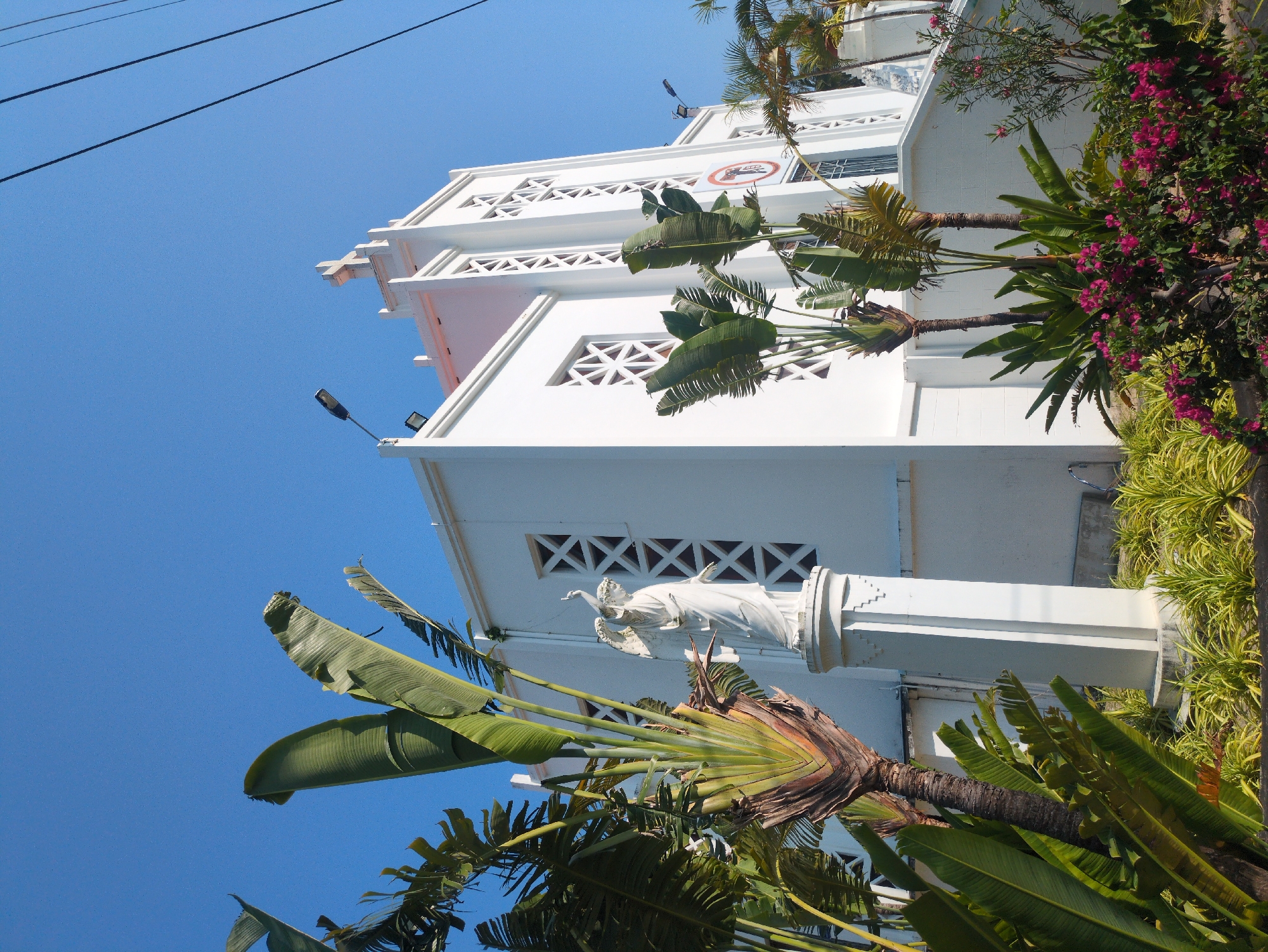 The image depicts a low-angle view of a white building with geometric architectural details, possibly a church or religious structure, in South Quy Nhon Ward, Vietnam. The structure is adorned with a white statue, possibly a religious figure. Lush tropical foliage, including banana trees and flowering plants, surrounds the building. Overhead power lines are visible against a clear blue sky.