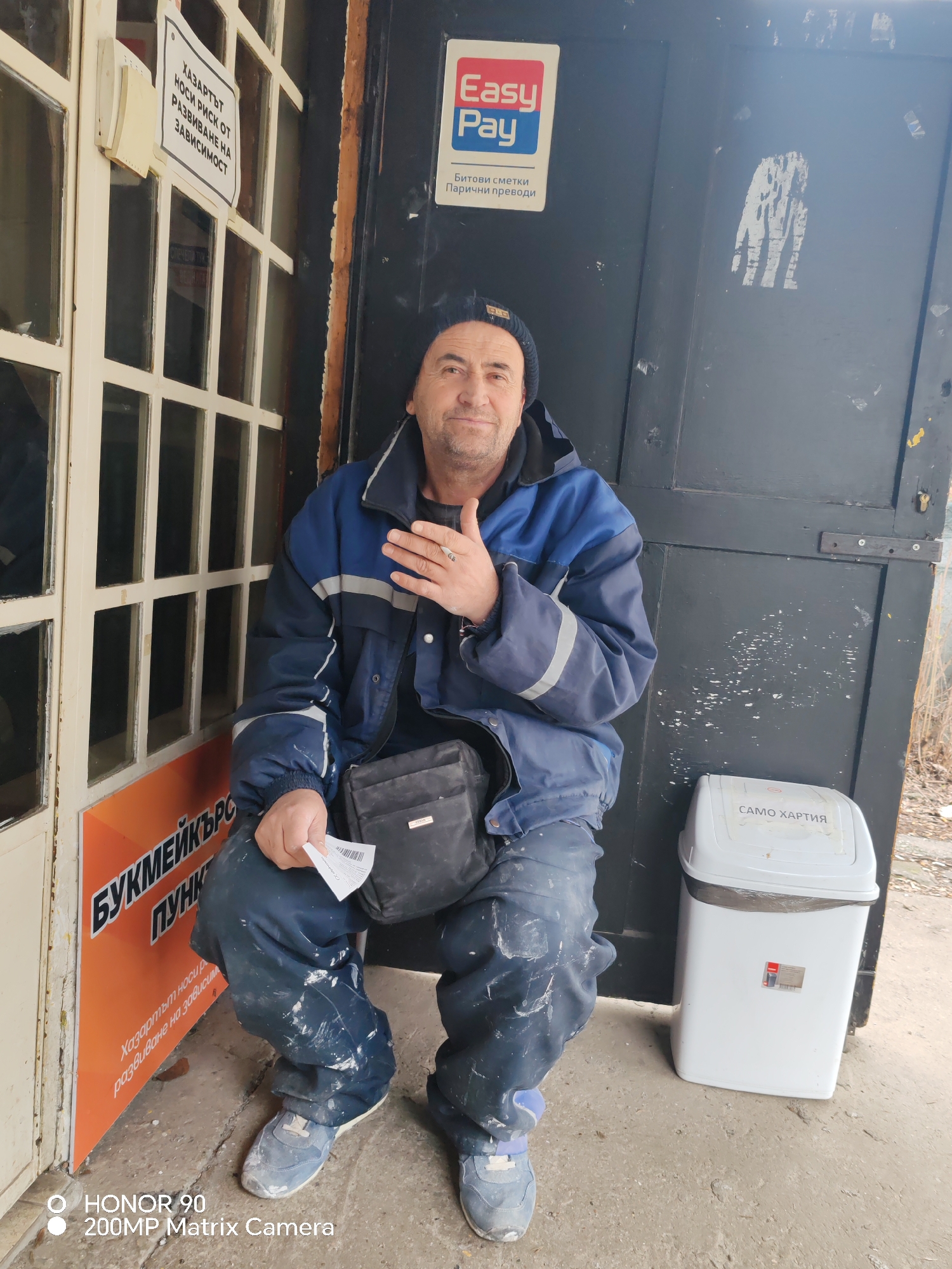 A man sits on a stoop outside a small building in Sofia, Bulgaria. He wears a blue jacket and pants splattered with paint, a dark knit cap, and carries a small black bag. In his hand, he holds a betting slip. An "EasyPay" sign is above him, and a sign on the door reads "Hazardous. Carries a risk of developing addiction." To his right, is a white trash can labeled "SAMO HARTIA" (Only Paper). A betting shop sign is to his left. The scene appears to be in a run-down or neglected area.