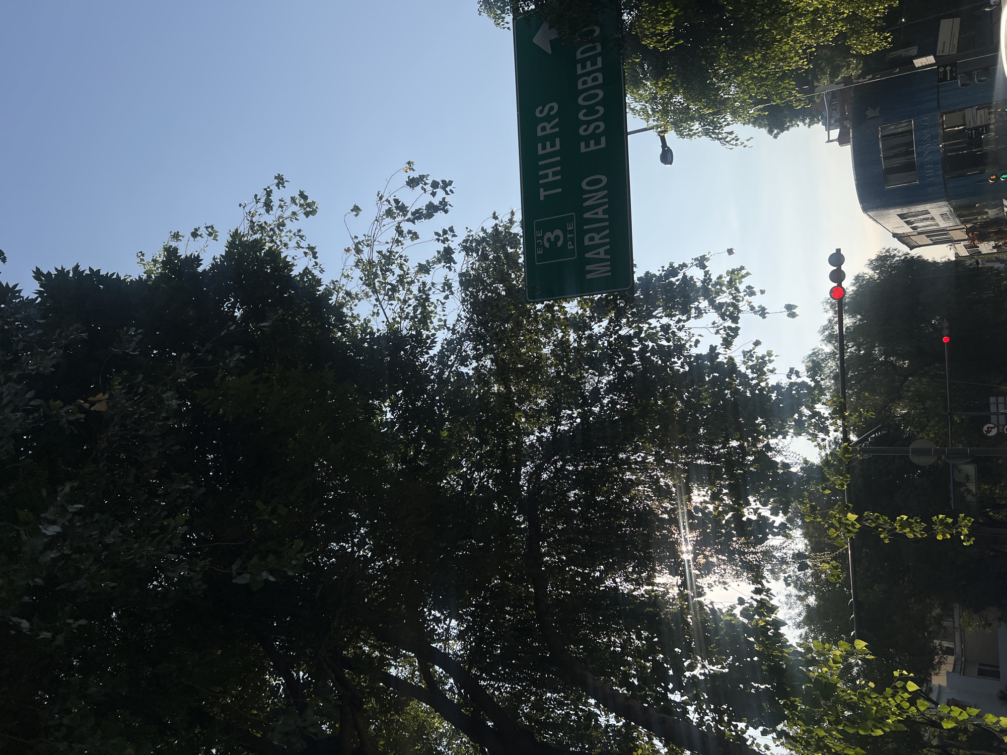 The image shows a street view in Mexico City, Mexico. A green street sign indicates directions to Thiers and Mariano Escobedo avenues. A traffic light is visible, displaying a red light. Trees are prominent in the foreground, partially obscuring the view. A building, possibly a business, is seen on the right side of the frame. The sky is bright, suggesting daytime.