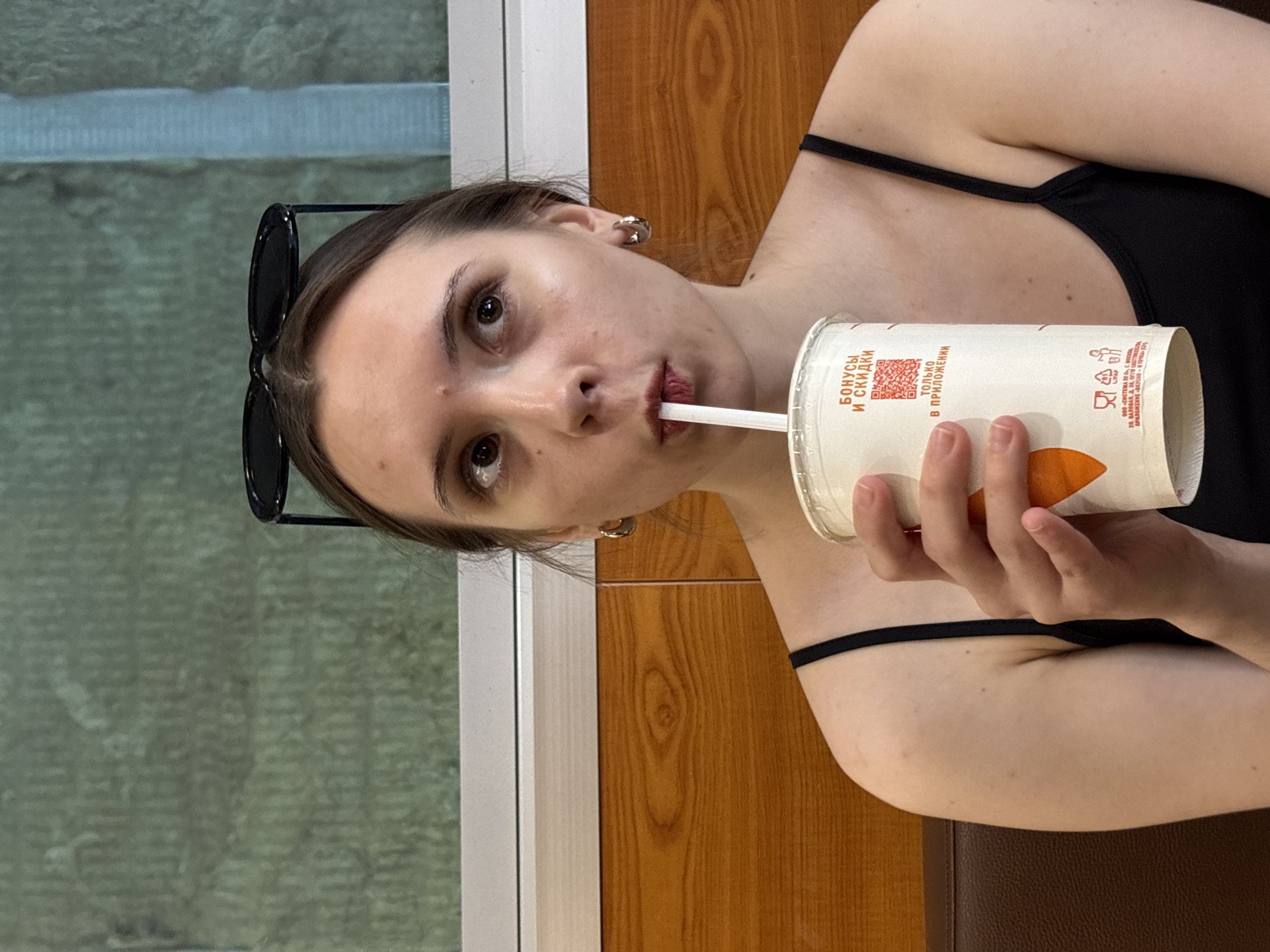 An image shows a young woman in an indoor setting, likely a cafe or fast-food restaurant in Saint Petersburg, Russia. The shot is a close-up portrait, oriented sideways, with the woman looking directly at the viewer. She has dark hair, styled away from her face, and is wearing a black top with thin straps. Black sunglasses are perched on top of her head.
The woman is actively drinking from a white disposable cup through a straw, with her lips pursed around the straw. Her expression is neutral and calm. Her right hand holds the cup. Visible text on the cup, in Russian, reads "БОНУСЫ И СКИДКИ" (BONUSES AND DISCOUNTS) and "ТОЛЬКО В ПРИЛОЖЕНИИ" (ONLY IN THE APPLICATION), along with details for "ООО «СИСТЕМА ПУ» О Г. МОСКВА" (SYSTEM PU LLC, Moscow) and the application "ВКУСНО-И ТОЧКА" (Vkusno-i Tochka), a prominent fast-food chain in Russia. An address for Valovaya Street in Moscow is also printed.
The background features a light brown wooden paneling to the right of the woman, and a blurred, textured green/grey surface, possibly an outdoor view or another part of the building through a window, to her left. She appears to be sitting on a dark brown, possibly leather or vinyl, seat. The lighting is soft and even, indicating an indoor daytime environment.