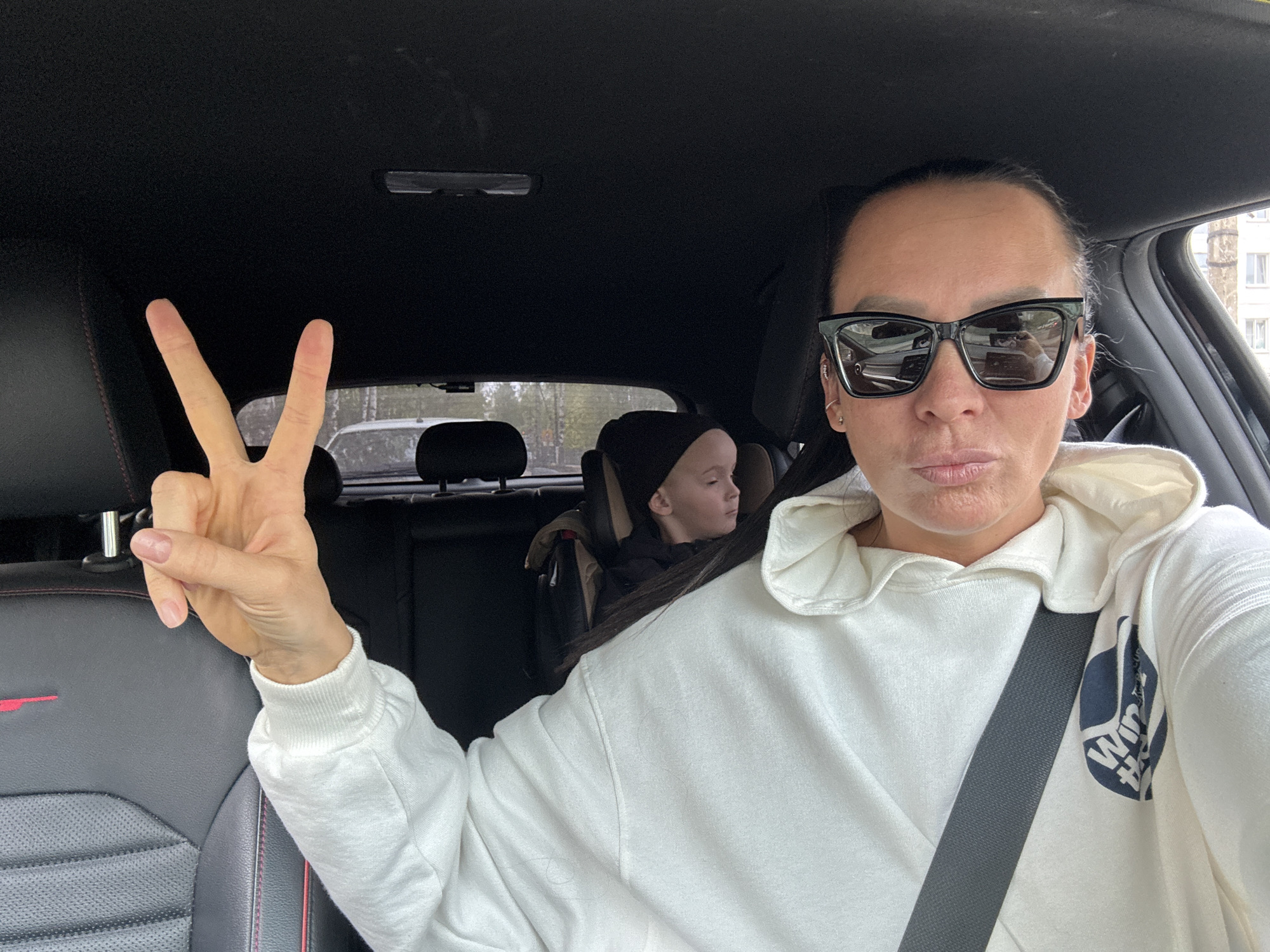 A woman is taking a selfie while sitting in the driver's seat of a car. She is wearing sunglasses and a white hoodie, and she is holding up two fingers in a peace sign. A child wearing a black hat is seated in the back of the car. The car's interior is black with red trim on the seats. Through the back window, trees and a building can be seen, suggesting an urban setting. The location is Chaikovsky, Russia, based on the availability of information about the image.