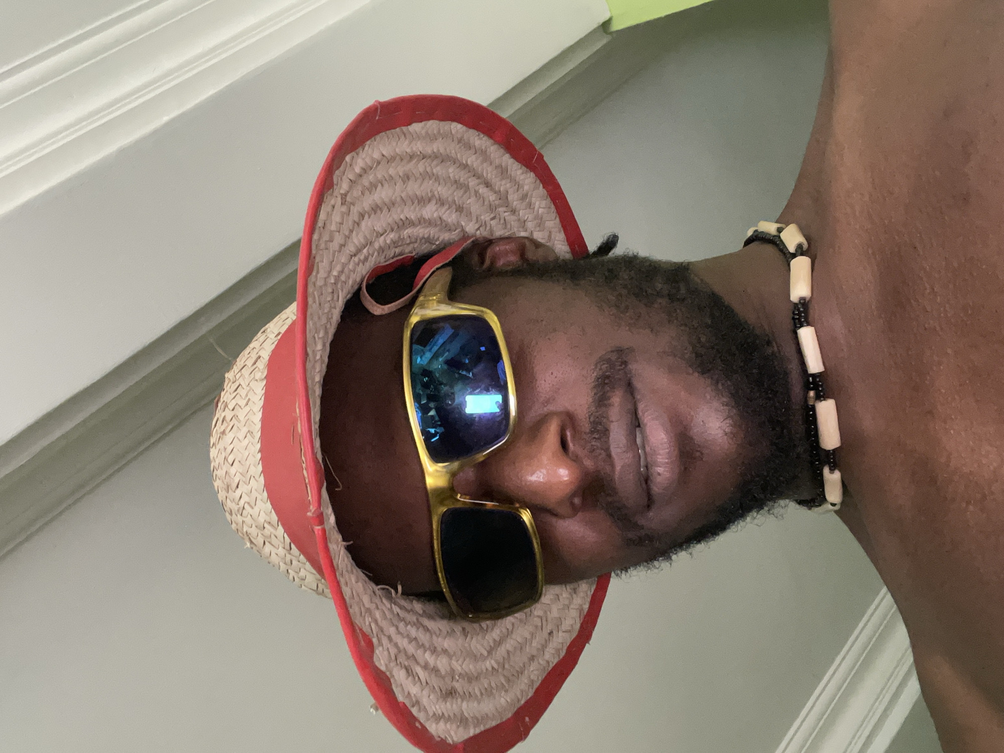 An African-American man is wearing a straw hat with a red brim and gold framed sunglasses. He is also wearing a beaded necklace. The location is difficult to determine without further visual cues or information, and I am unable to discern any details that would suggest it is in the city of Abuja, Nigeria.