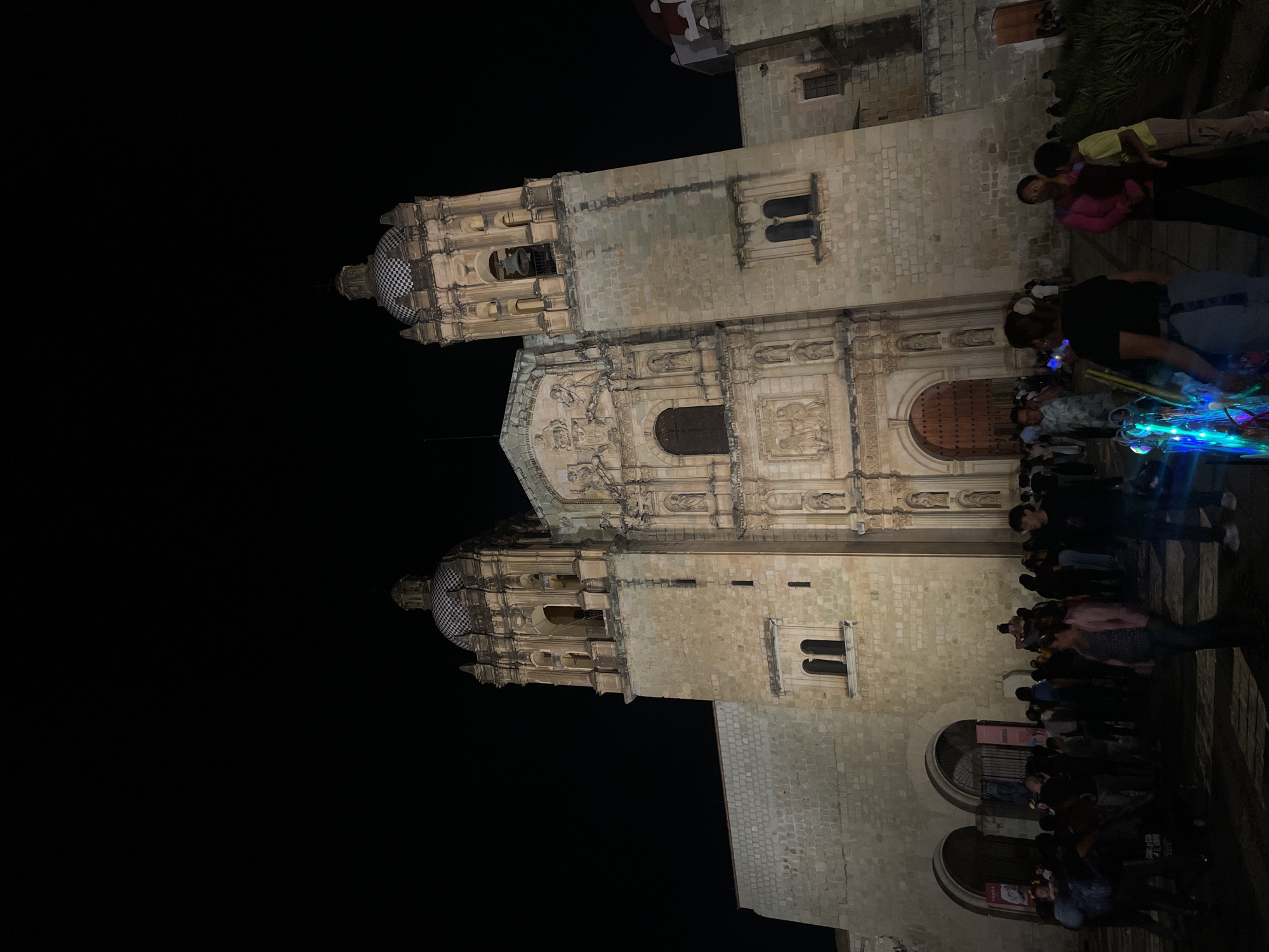 At night, the lit Oaxaca Cathedral in Oaxaca City, Mexico, stands tall with its baroque facade and tiled domes, with a crowd of people milling about near its entrance. Several individuals are standing in the foreground, with some possibly engaging in conversation. A colorful light display or structure is visible in the crowd.