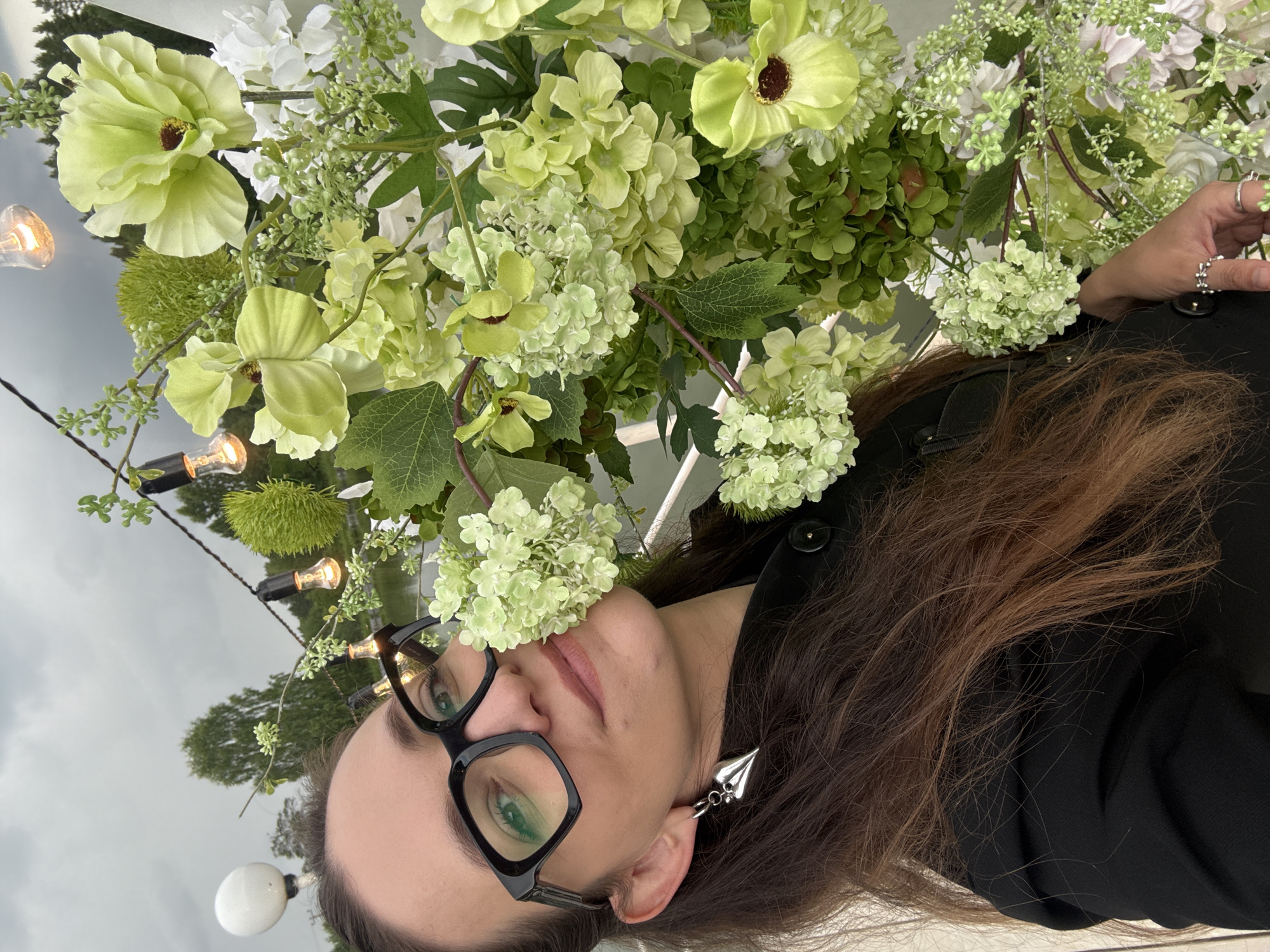 A woman with long brown hair and black-rimmed glasses is pictured up close, partially obscured by a large floral arrangement. She is wearing a black jacket and a silver earring shaped like a heart. The floral arrangement is a mix of light green and white flowers, some of which are large and open, while others are small and clustered. Behind the woman and the flowers, a cloudy sky can be seen, along with trees and a body of water, suggesting an outdoor setting. String lights with exposed bulbs are also visible, adding to the ambiance. The scene appears to be a decorative setup, possibly for an event. There is no visible indication of the specific city of Votkinsk, Russia within the image itself.