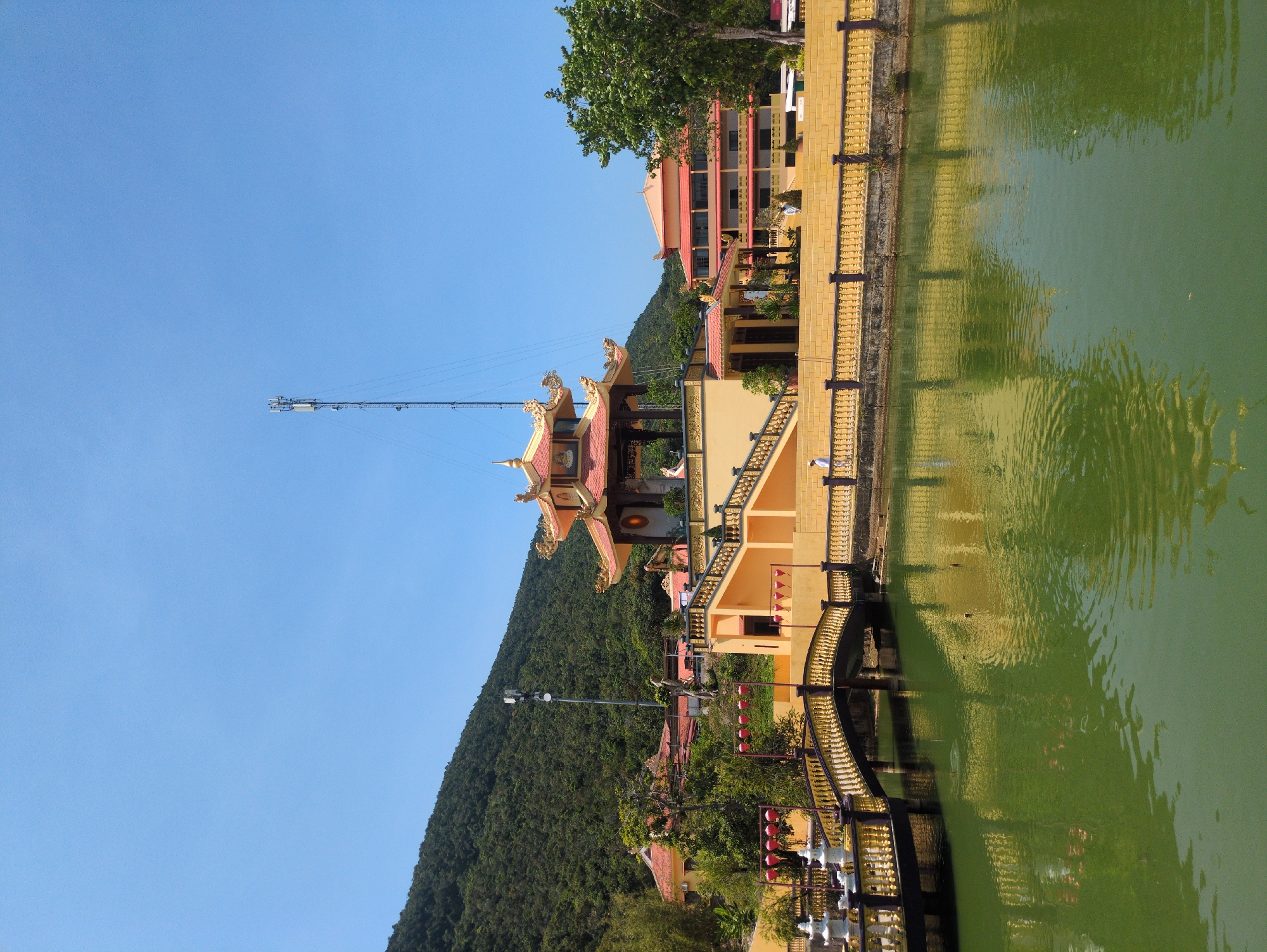 The image captures a view of Hùng Long Pagoda, a Buddhist temple in Phú Quốc, Vietnam. The scene includes the pagoda's multi-tiered roof structure with decorative dragon figurines, a bridge leading to the temple across a green-tinted pond reflecting the buildings, and a mountain densely covered with trees in the background. A tall communication tower stands near the temple, contrasting with the traditional architecture. The setting is under a clear blue sky, indicating daylight. Architectural details include yellow walls and staircases, and red lanterns are hung around the buildings. Vegetation is abundant around the temple grounds.