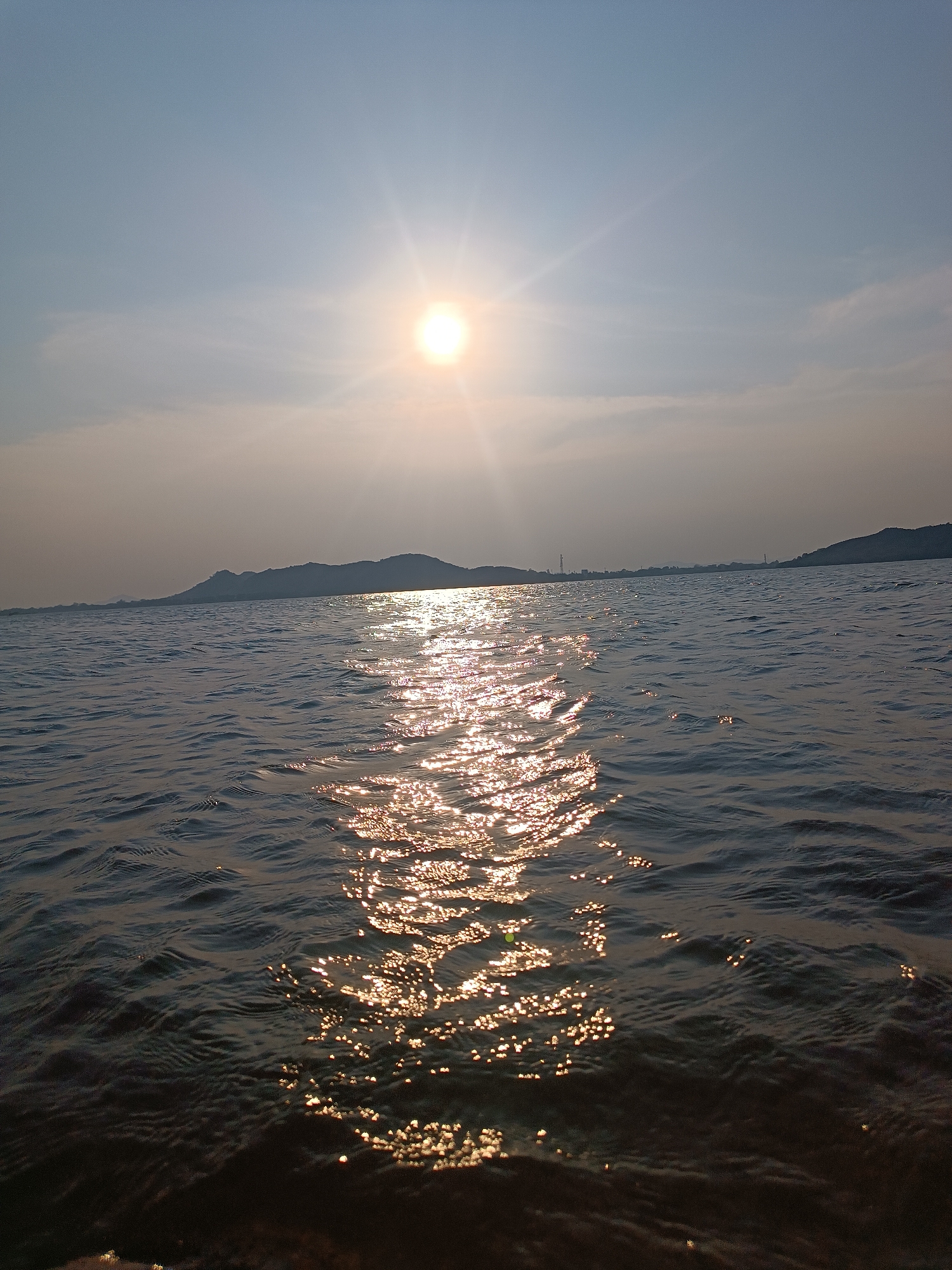 The image depicts a body of water with the sun reflecting off its surface, creating a shimmering effect. In the background, there are hills and possibly some distant structures, suggesting a landmass across the water. The location is likely in or near Kammavaripalli, India, due to the geotagged information. The scene lacks specific details about people, actions, or interactions. The environment is characterized by the presence of water, distant land, and sunlight.