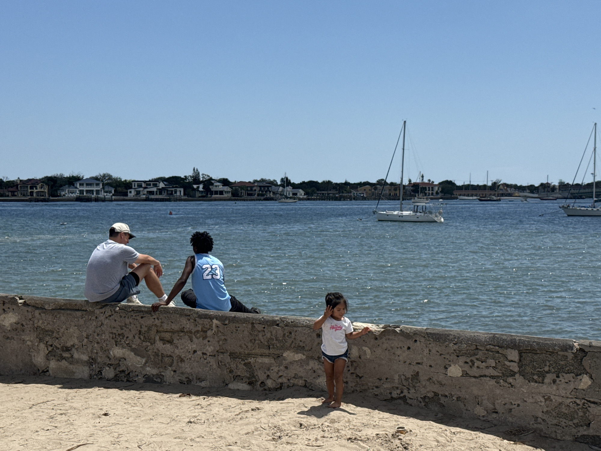 This outdoor medium shot depicts a sunny day by a waterway in Saint Augustine, United States of America. In the foreground, a sandy area leads up to a weathered, grey concrete wall. A young child, appearing to be a girl with dark hair, stands barefoot in the sand near the wall, wearing a white t-shirt with "Happy" partially visible text and blue shorts. She is looking towards the water or the individuals seated on the wall.
Two adults are seated on the concrete wall, facing away from the viewer and looking out over the water. One individual wears a light grey t-shirt, dark shorts, and a light-colored baseball cap. Beside them, another person wears a light blue jersey with the number "23" visible on the back, along with dark pants.
The midground features a wide expanse of sparkling blue water with small ripples. Several sailboats and other smaller vessels are dotted across the water. In the background, the opposite shoreline is visible, lined with a variety of houses and buildings, interspersed with green trees. The sky above is clear and bright blue, indicative of a pleasant, sunny day. The scene conveys a relaxed, leisurely atmosphere by the water.