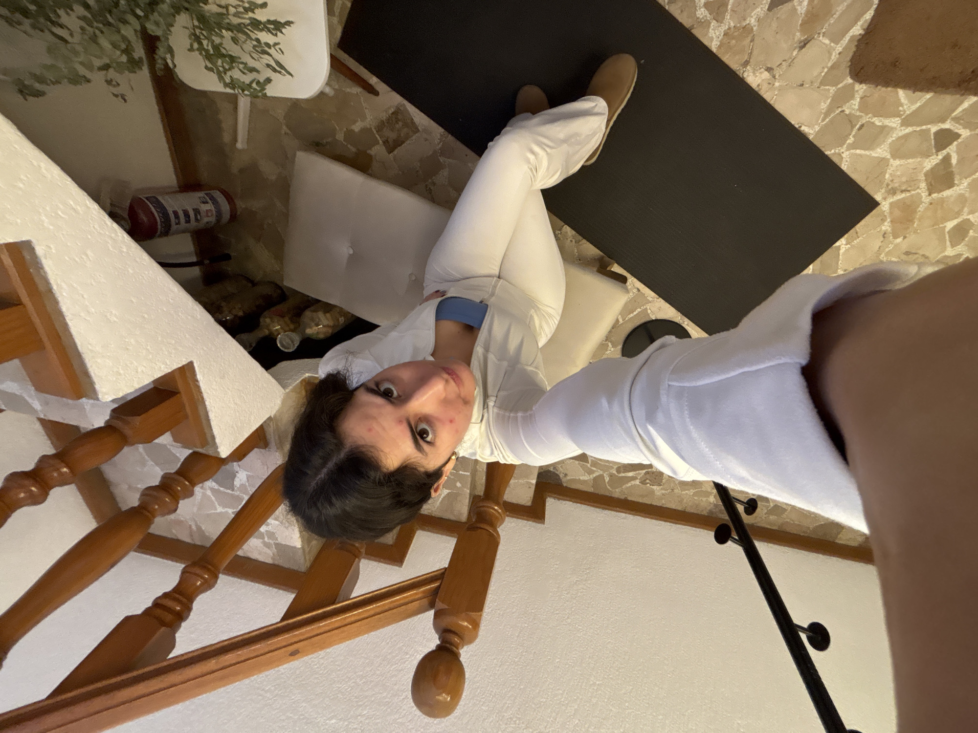 A woman in a white outfit and brown shoes is sitting on a white cushion placed on a floor with mosaic tiles. A black yoga mat is nearby. To the left, a wooden staircase is visible. In the background, there is a fire extinguisher and what appears to be a plant. The location is Puebla, Mexico.