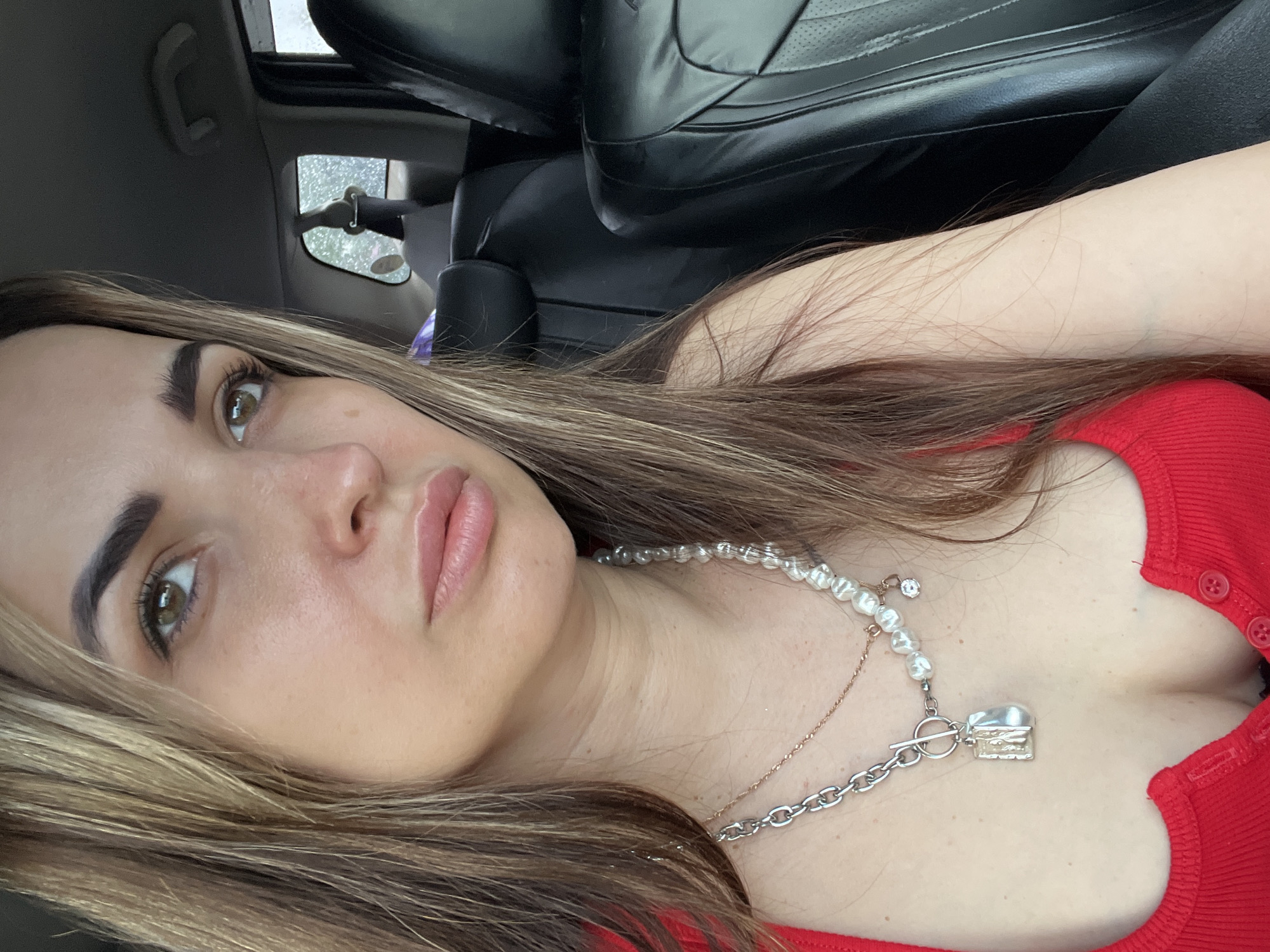 A woman is depicted in a close-up, slightly tilted portrait taken inside a car. Her head and upper torso fill the frame, positioned as if looking upwards and slightly to the side. She has long, dark brown hair with lighter highlights, which cascades around her face and over her shoulders. Her left eye is visible, featuring a brown iris and well-defined eyelashes, complemented by thick, dark eyebrows. Her full lips are slightly parted, showcasing a natural pink hue. Light freckles are visible across her nose and cheeks.
She is wearing a bright red ribbed top with small matching buttons running down the front. Around her neck, she sports a layered necklace set. One strand consists of irregular white pearls, while another is a delicate gold chain. A prominent silver chain with a T-bar clasp and a chunky, textured silver pendant completes the ensemble.
The background reveals the interior of a vehicle, with plush black leather seats visible behind her. Through a car window in the upper left, raindrops are distinctly visible, indicating rainy or recently rainy weather outside. The overall lighting suggests it is daytime. Her expression is calm and somewhat contemplative, with her gaze directed slightly away from the camera. There is no visible text in the image.