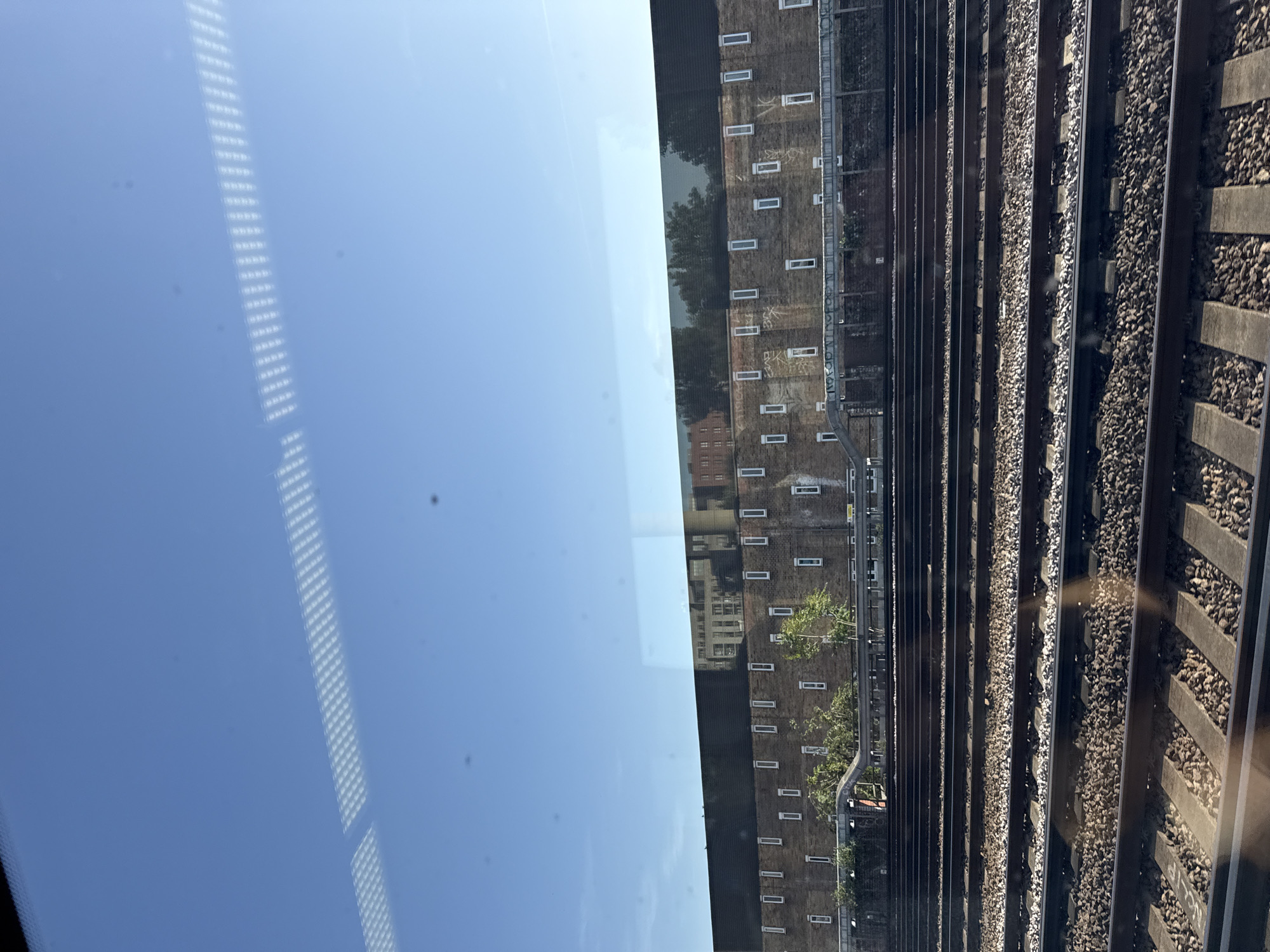 The image shows a view from a train window, depicting a railway track, a brick building, and a reflection of the sky. The railway track is composed of multiple parallel tracks with gravel and concrete sleepers. The brick building runs parallel to the tracks, with a series of small, evenly spaced windows. The sky, reflected in the train window, is clear and blue. The location appears to be urban.