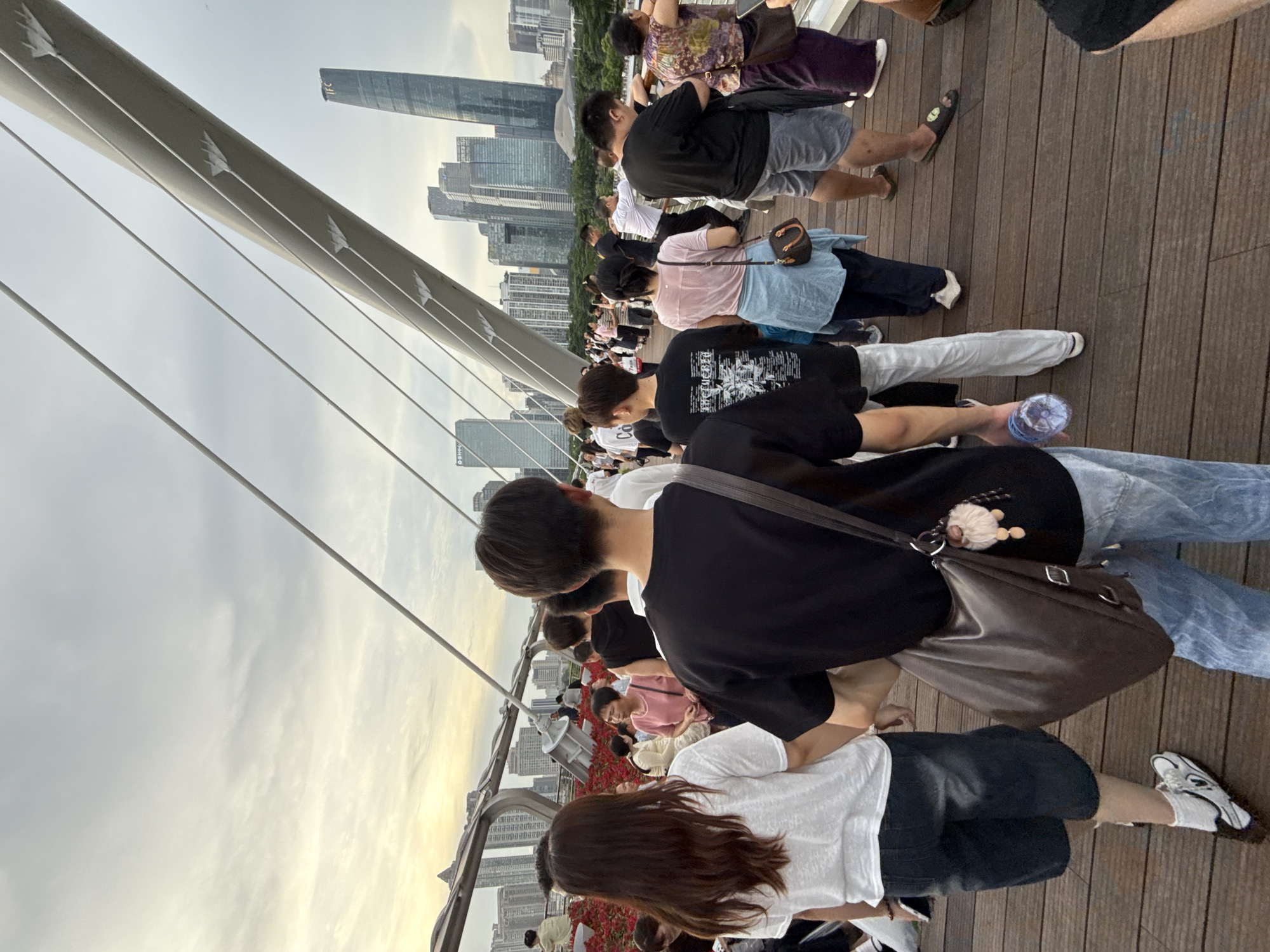 The image depicts a group of people standing on a wooden deck, possibly a bridge or boardwalk, in an urban setting. The subjects are a mix of individuals, including men and women of various ages, dressed casually. They are oriented in different directions, with many looking towards a distant cityscape. The scene is characterized by a large, angled bridge structure with cables, suggesting it's part of a modern architectural design. In the background, a cluster of tall buildings, including skyscrapers, indicates an urban environment.
The time of day appears to be late afternoon or early evening, as the sky is overcast with soft light, and hints of a sunset glow are visible. The weather seems to be cloudy, but not actively raining. The people are standing and observing, some are walking. The atmosphere seems relaxed and observational, with no overt signs of urgency or distress.
A few notable details include a brown shoulder bag with a fluffy keychain, a plastic water bottle, and a pair of white athletic shoes. The wooden planks of the deck are clearly visible, arranged lengthwise. The architecture of the bridge and the distant skyscrapers are prominent features of the setting. The image is taken from a slightly elevated perspective, looking down on some of the people and across towards the city. There is no visible text in the image.