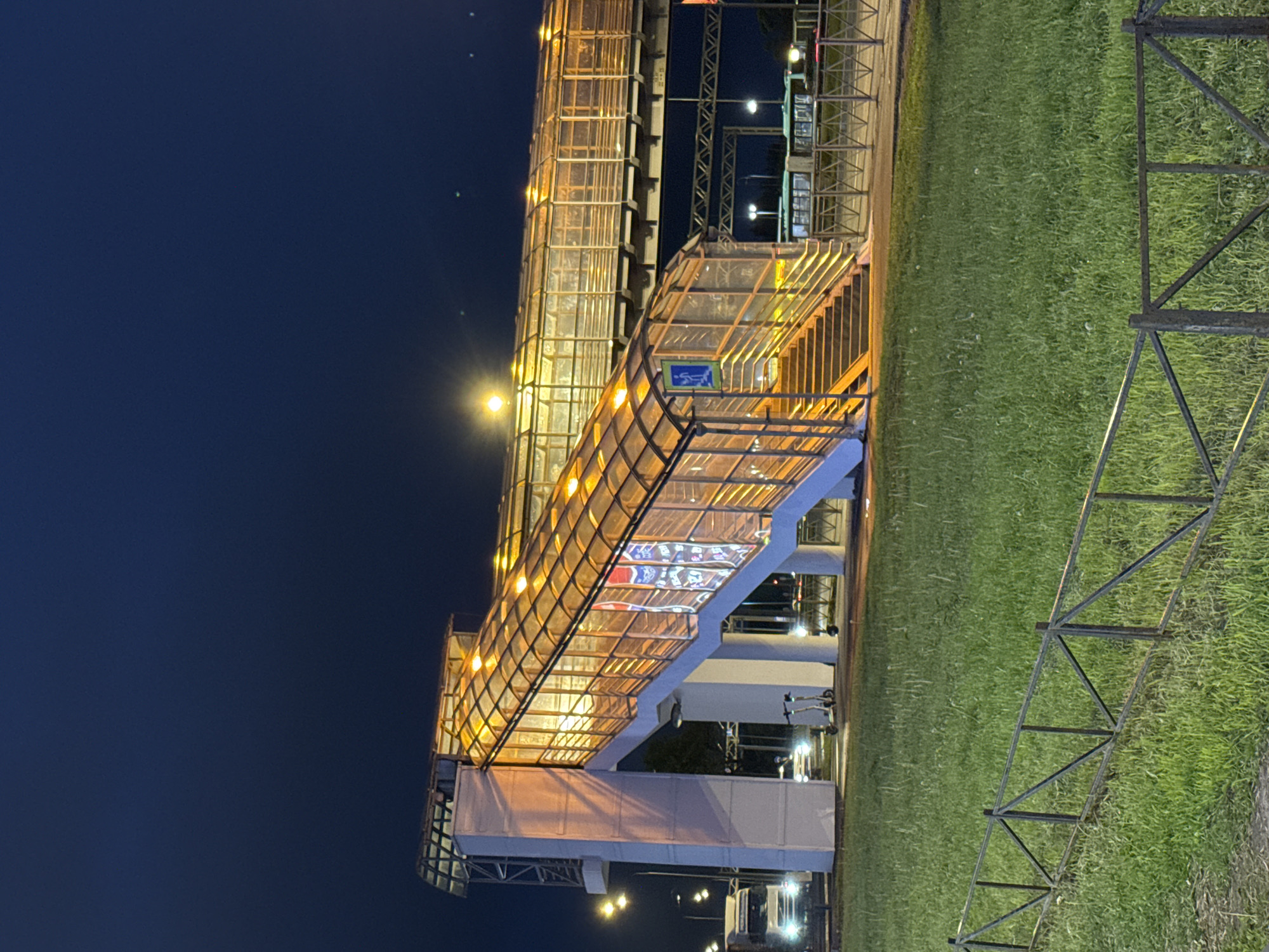 The image depicts an elevated pedestrian bridge at night in Saint Petersburg, Russia. The bridge is enclosed with glass and illuminated from within, casting a golden glow. It crosses over an area with grass and a metallic structure. The sky is dark. There are no people visible in the image.