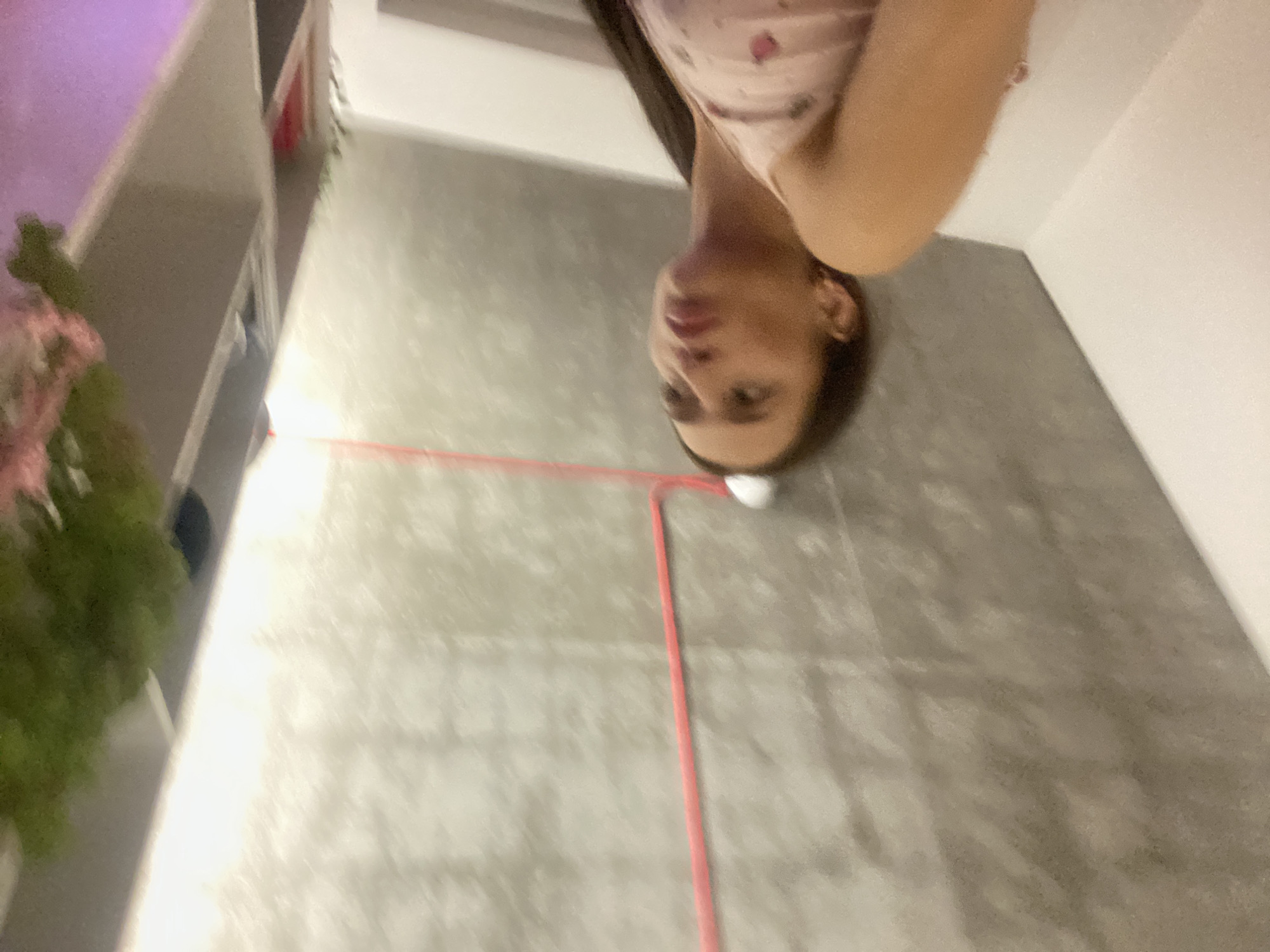 The image shows a young woman with long dark hair, wearing a pink tank top with a strawberry pattern. She is looking upwards and to her right with her mouth slightly pursed. She is in an indoor environment, possibly an apartment, with a concrete ceiling and a white wall. A bright pink cable runs across the ceiling. To the right of the woman, a shelf is visible with some greenery and pink flowers. The lighting is bright, with a strong light source coming from the right. The image appears to be taken at a low angle. There is no indication of the specific location in London, United Kingdom.