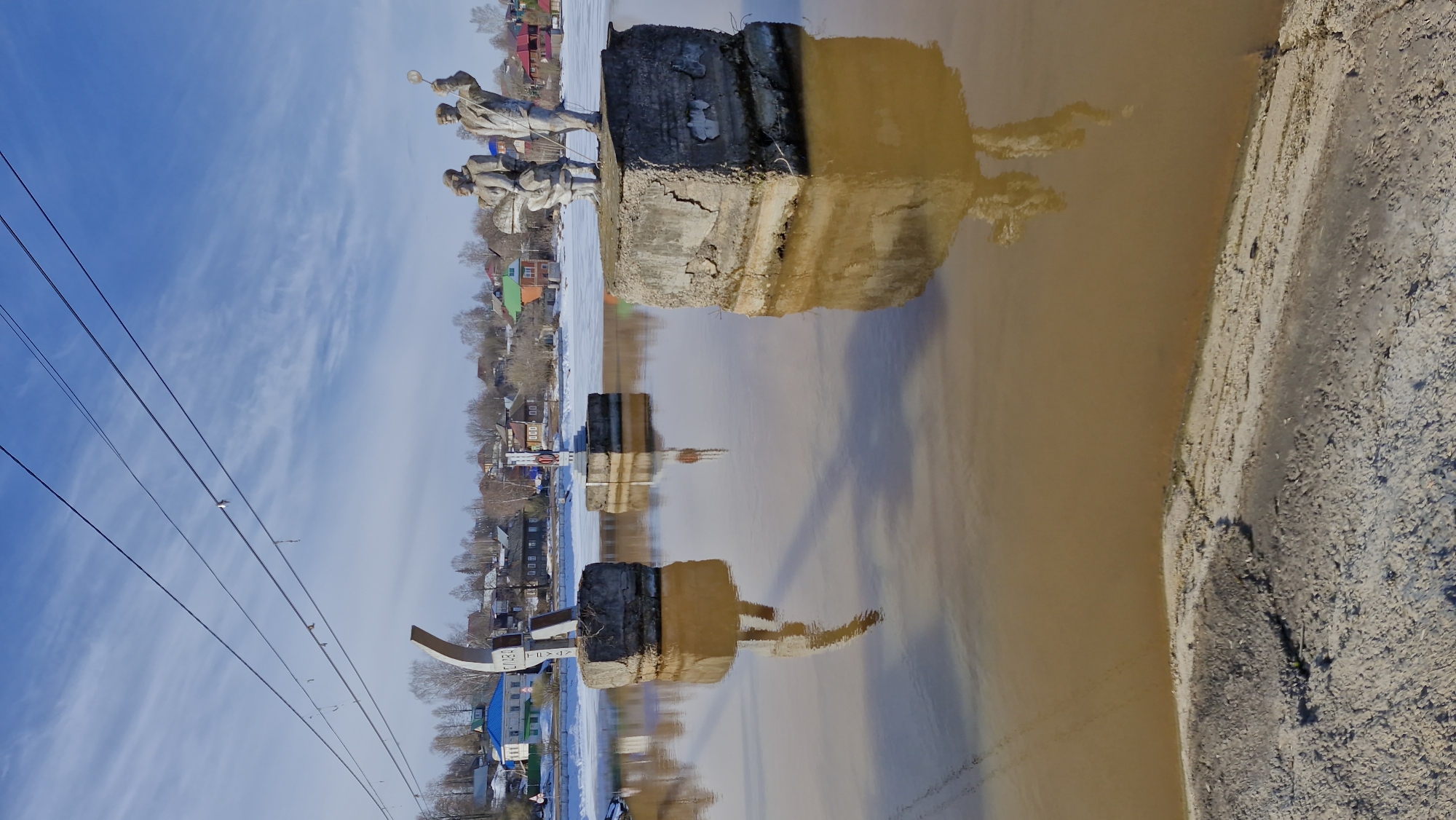 The image depicts a reflective body of water, possibly a river, with visible support structures or concrete blocks extending from the water's edge. These structures are situated near the city of Otchor, Russia, as indicated by the surrounding buildings and infrastructure. At least one statue can be seen above the water line on the left side of the frame, with its reflection visible in the water. There is also evidence of power lines overhead, suggesting an urban or semi-urban location. The surrounding environment includes buildings, bare trees, and indications of a seasonal thaw or flood.