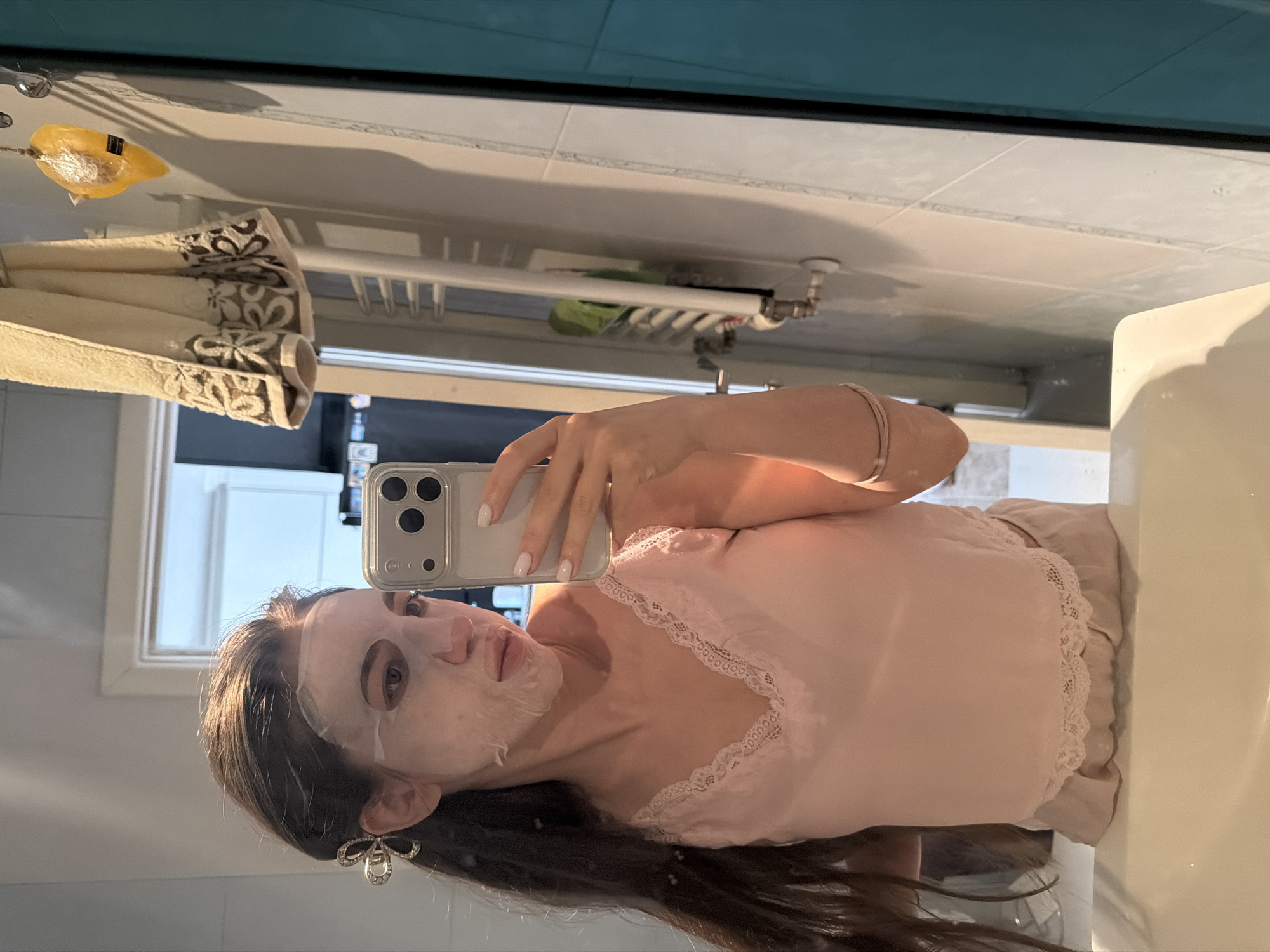 In a brightly lit bathroom, a young woman with long, dark hair pulled back with a sparkly bow, is taking a selfie in a mirror. She is wearing a light pink camisole with delicate lace trim. Her face is covered with a white sheet mask. She is holding a smartphone in her right hand, capturing her reflection. The bathroom appears to be in a modern apartment, with tiled walls and a sink visible in the foreground. A towel hangs on a rack in the background, along with a yellow, soap-like object. The overall impression is one of personal grooming and self-care in a domestic setting. The image does not provide any specific clues about its location in Sofia, Bulgaria.
